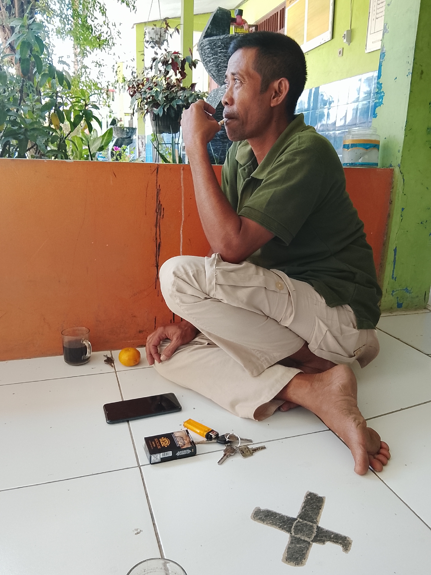 A middle-aged man is sitting on the floor outside a building, smoking a cigarette. He is wearing a green polo shirt and light-colored pants. On the floor in front of him are a glass of coffee, a small orange, a smartphone, a pack of cigarettes, a lighter, and a set of keys. Behind him is an orange wall and a green wall with some writing on it. The floor is tiled. The scene appears to be in Kebasen, Indonesia.