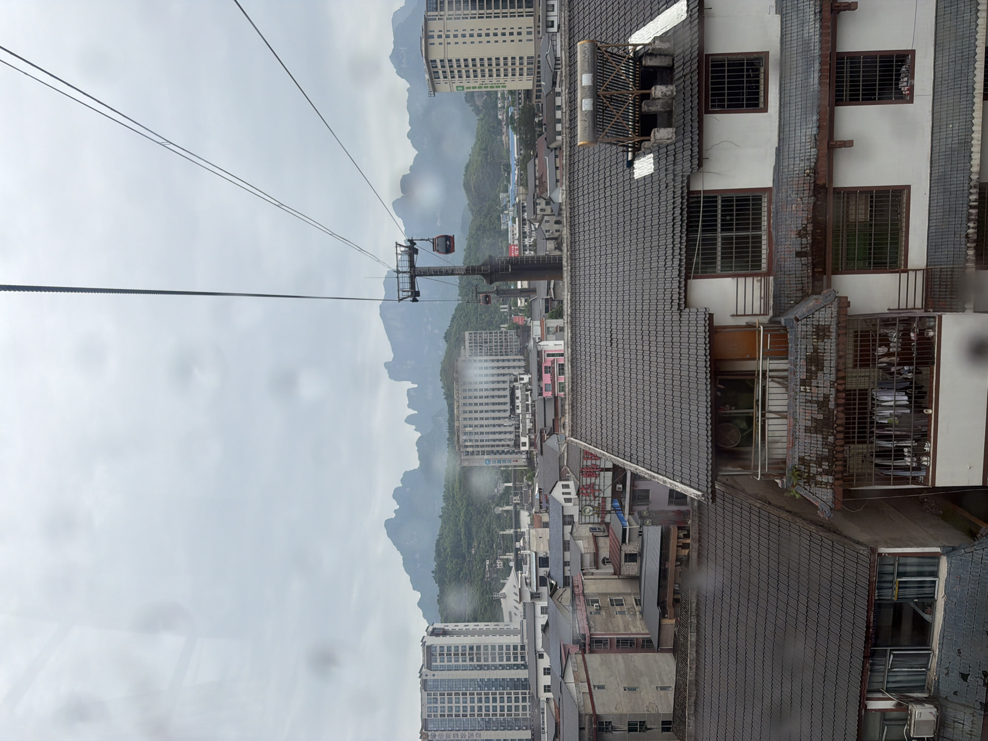 From an elevated vantage point, the image captures a view in Zhangjiajie, China, encompassing a blend of urban and natural elements.
In the midground, a city landscape is visible, composed of varied structures and building heights. Dominating the foreground, the rooftops of adjacent buildings are visible, some adorned with satellite dishes. A cable car system intersects the view, with cabins suspended along cables, suggesting a tourist attraction or transport route.
In the background, karst mountains rise through a misty haze, indicative of the unique geological formations for which the Zhangjiajie area is known. The overall atmosphere hints at cloudy or overcast conditions.
