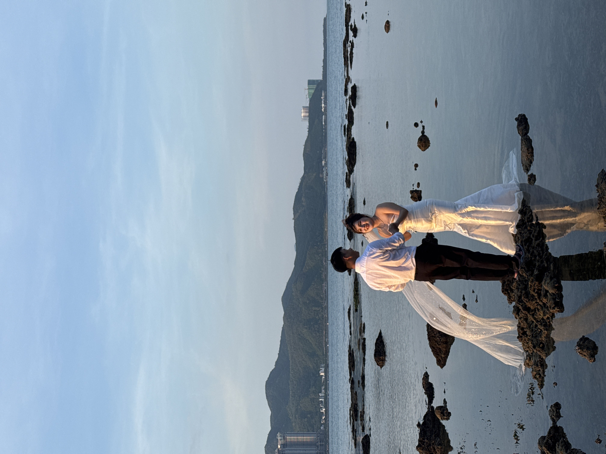 In Bac Nha Trang Ward, Vietnam, a newlywed couple is captured in a romantic moment on a rocky shoreline during what appears to be a sunset. The bride, dressed in a form-fitting white wedding gown with a mermaid silhouette, stands facing her groom, her hands clasped at her chest and a joyful smile on her face. The groom, in a white collared shirt and black pants, stands opposite her, his gaze directed towards his bride. A sheer, sparkling veil flows from the bride, extending towards the groom and creating a dynamic visual element in the scene. They are positioned on dark, irregular rocks emerging from the shallow, reflective water. In the background, the tranquil sea stretches towards a distant, mountainous coastline dotted with buildings, including a tall, modern high-rise structure on the left and construction cranes on the right. The sky above is a clear, soft blue with faint wisps of clouds, indicating the serene end of the day. The overall atmosphere is peaceful and celebratory, suggesting a wedding photoshoot or a moment of post-nuptial bliss.