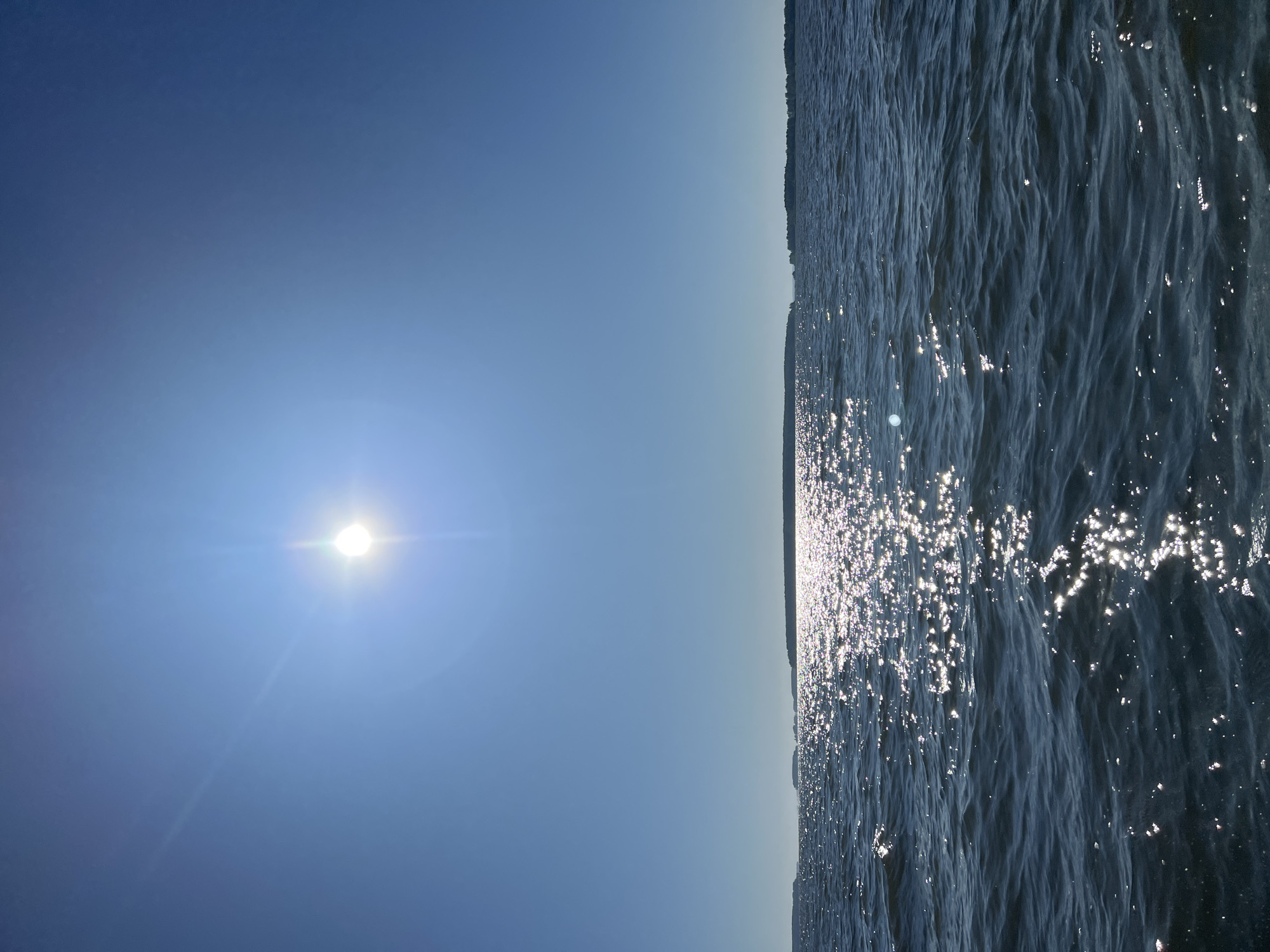The image depicts a bright, sunny day over a body of water, likely a lake or sea, with a faint landmass on the horizon. The sun is high in the clear blue sky, creating a strong glare on the rippling water's surface. The water itself is dark blue and appears choppy. The horizon is marked by a dark, silhouetted line of trees or land. No people, structures, or any signs of urban environment are visible. The scene suggests a natural, open-air setting. The location is not specifically identifiable as Novopichugovo, Russia, from the visual information provided.