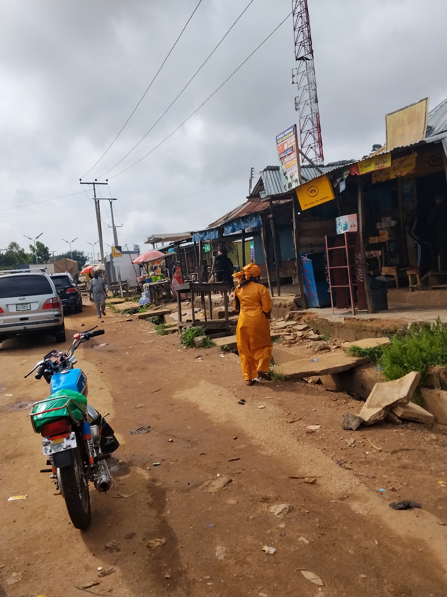 This is a wide-angle, eye-level, exterior shot of a dusty street scene in Kuru Dungus, Nigeria. The sky is overcast and a telecommunications tower is visible in the background. On the right side of the street, several small market stalls with corrugated metal roofs are set up. A woman in an orange outfit is walking away from the camera past these stalls. In the foreground, a blue and green motorcycle is parked. The ground is a mix of dirt and gravel, with some debris scattered around. A silver SUV and a black car are parked on the left side of the street, and a man in a gray shirt is walking on the street between the motorcycle and the parked cars. There are several utility poles and wires throughout the scene.