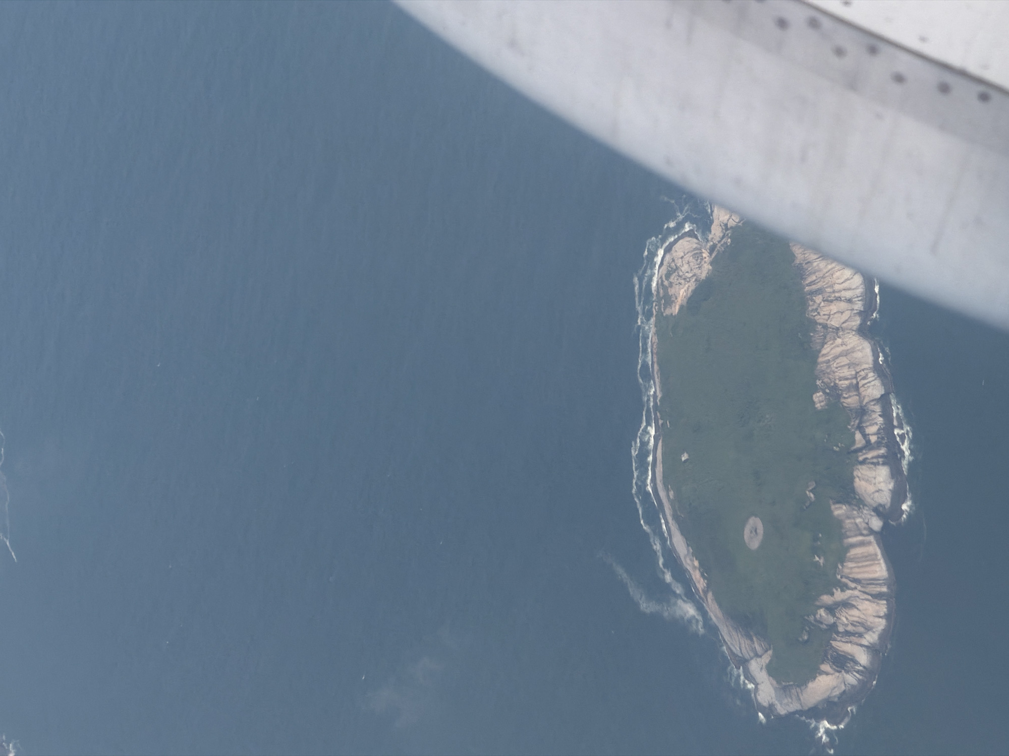 This is an aerial photograph of an island in the ocean, viewed from an aircraft. A portion of the aircraft's wing or fuselage is visible in the foreground on the right side of the image. The island is oval-shaped and covered with green vegetation. The coastline of the island is rocky, with waves crashing against it. The surrounding water is dark blue. The image does not provide enough detail to identify the specific location in Hong Kong, China.
