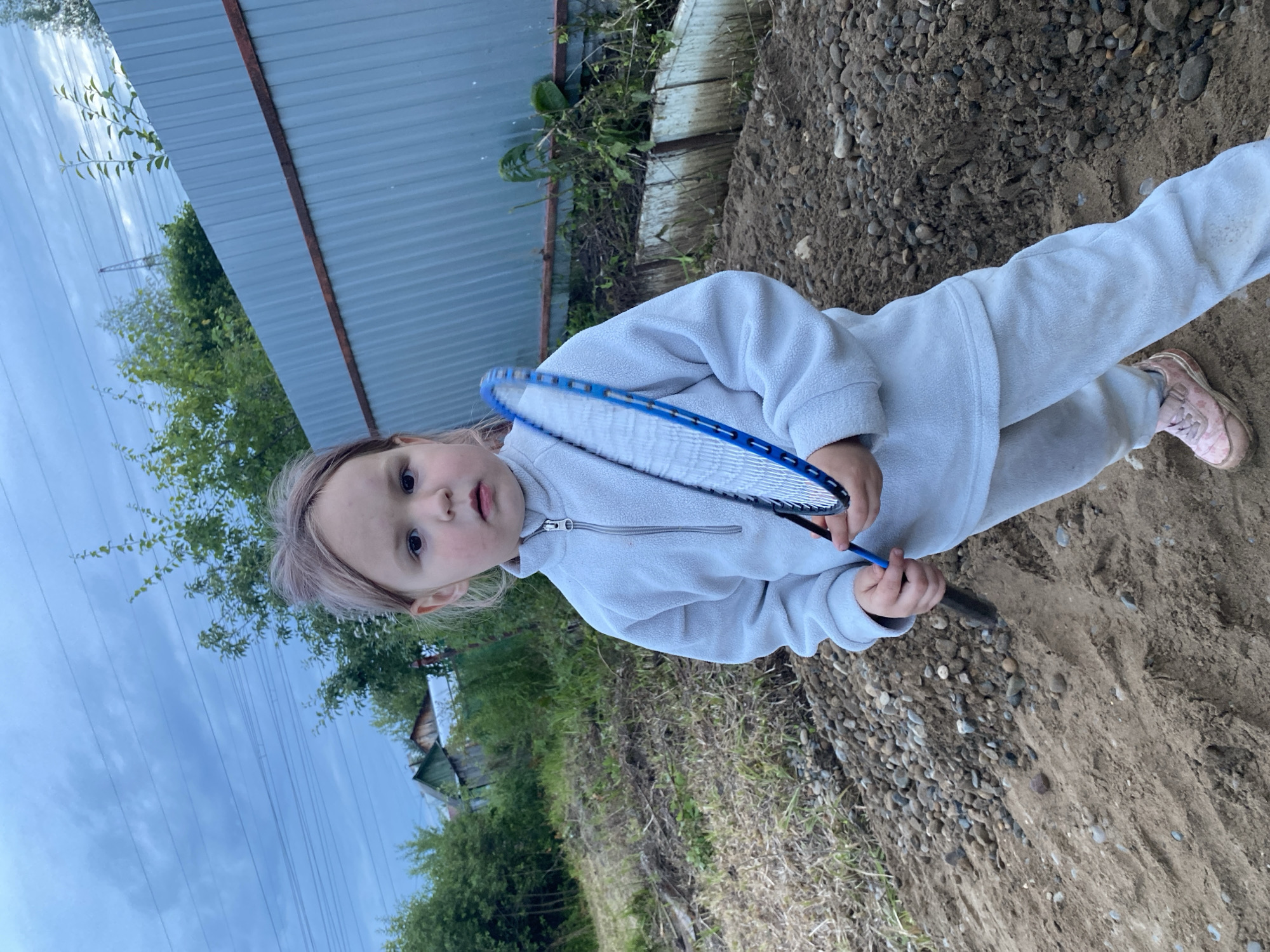 A young girl, likely a toddler, is pictured outdoors holding a blue badminton racket. She is wearing a light grey fleece tracksuit and pink sneakers. Her hair is light brown with some lighter streaks, and she has a curious expression on her face. The girl is standing on sandy ground mixed with small pebbles.
Behind her, there is a grey metal fence and overgrown vegetation. In the background, a wooden structure, possibly a shed or part of a house, is visible amongst trees. The sky is overcast, suggesting a cloudy day. The image is taken at ground level, looking up slightly at the girl. The overall scene appears to be a rustic, outdoor setting, possibly a backyard or a rural area. The photograph is oriented vertically, with the girl positioned in the lower right quadrant of the frame.
