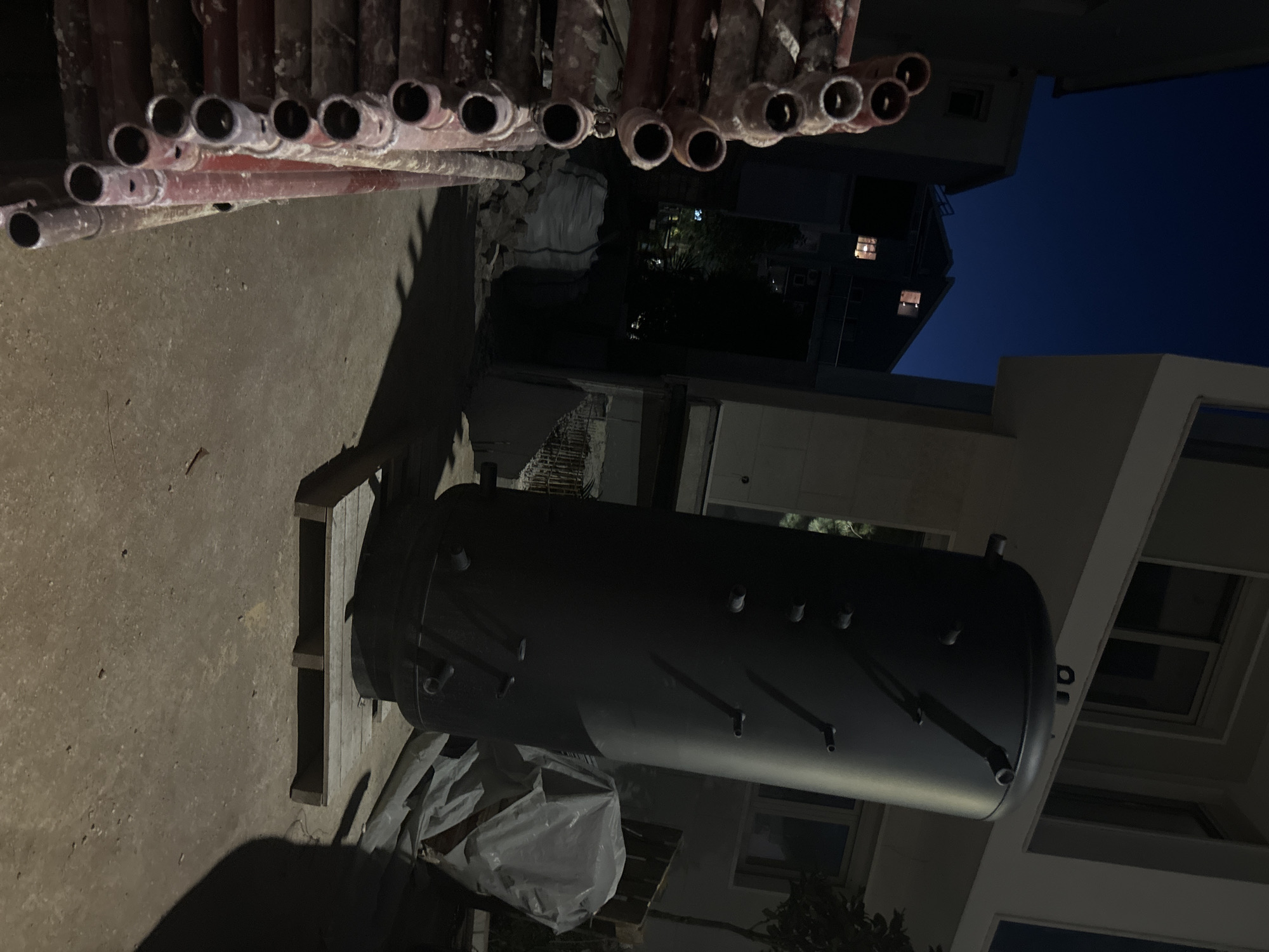 In Budva, Montenegro, a large black cylindrical water heater stands vertically on a wooden pallet outdoors at night. The water heater has several protrusions on its surface. To the left of the water heater, a stack of red scaffolding pipes is arranged. In the background, a modern building with windows is visible. The scene is illuminated by artificial light sources.