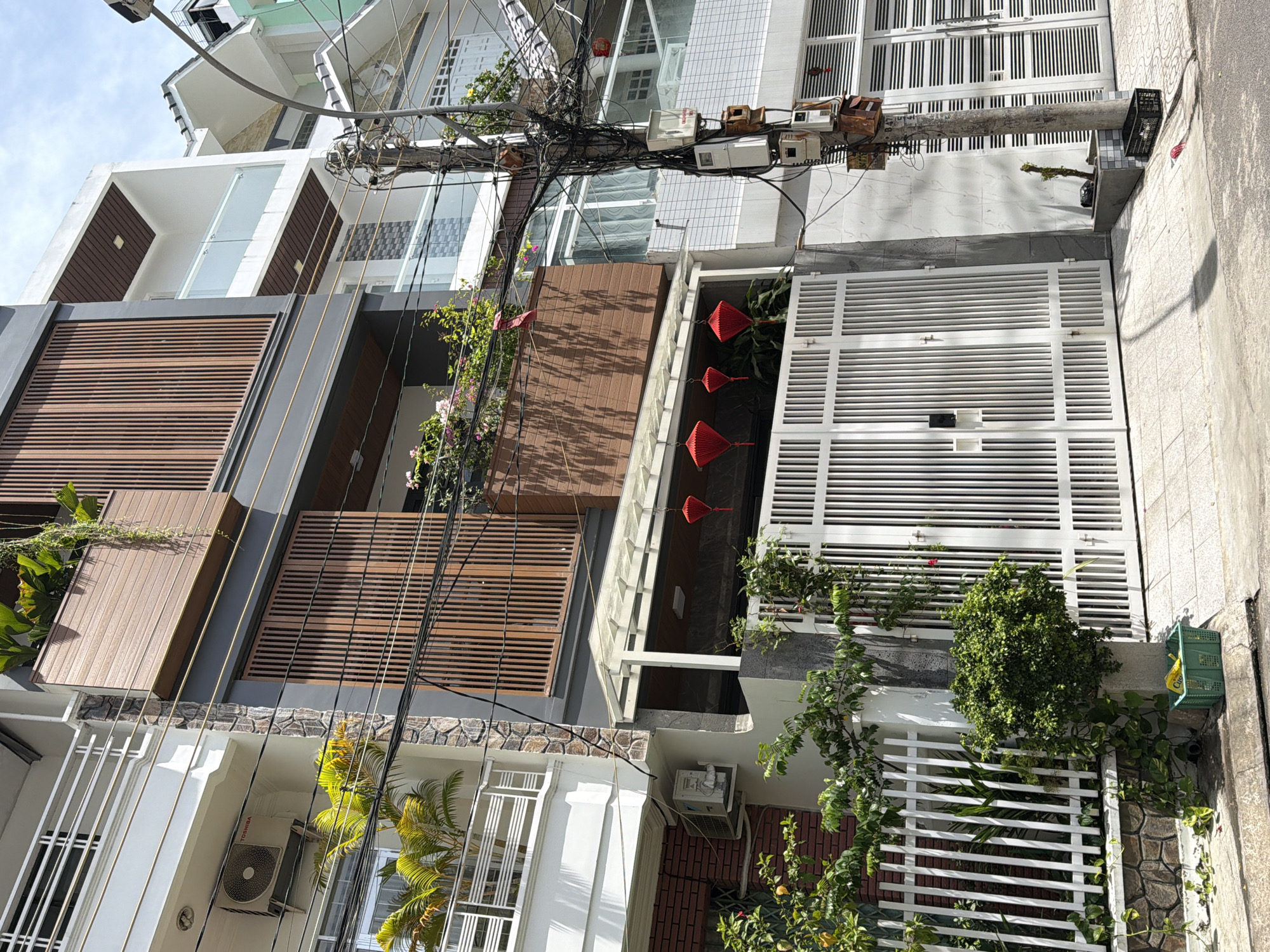 The image shows a street scene in Nha Trang Ward, Vietnam, focusing on the facade of a multi-story building. The building features a mix of modern architectural elements, including dark grey and white siding, wood-paneled balconies, and white railings. A large, white metal gate with vertical bars is prominent in the foreground, partially obscuring a paved walkway and the street. Above the gate, red paper lanterns are suspended.
Several potted plants and trees are visible, with lush green foliage cascading from balconies and a small bush near the gate. An air conditioning unit is mounted on the exterior wall of an adjacent, lower building to the left. Electrical wiring is densely networked around a utility pole situated to the right of the gate, with various electrical boxes attached. A green plastic crate containing yellow items is placed on the pavement near the base of the utility pole. The scene appears to be captured during daylight with a bright sky overhead.