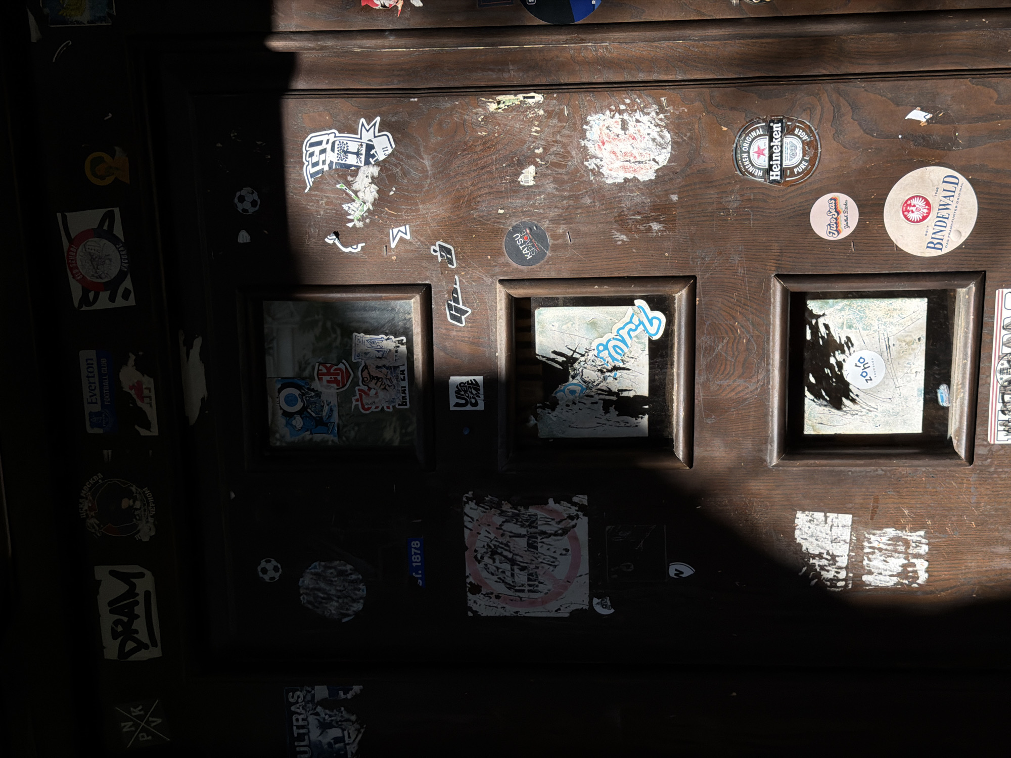 This image shows a close-up of a dark wooden door covered in various stickers. The door appears to be old and weathered, with visible wood grain and some wear and tear. Sunlight casts a dramatic shadow across the left side of the door, obscuring some of the stickers and details.
The stickers are varied in size, shape, and content. Some are faded and peeling, while others appear newer. There are stickers with text, logos, and what appear to be graphical designs. Among the visible stickers are what look like sports team logos (possibly football related), branded beverages (Heineken, Bindewald), and other assorted graphics. Some of the stickers are on individual panels of the door, suggesting the door is segmented into sections. The overall impression is that of a frequently visited or public-facing door that has accumulated a history of stickers over time.
The location is identified as Sabail Raion, Azerbaijan. While the image itself does not contain explicit markers of this location, the context provided suggests this is the setting. The specific environment is the exterior of a building, indicated by the door, and the presence of sunlight and shadows. There are no people or animals visible in the image.