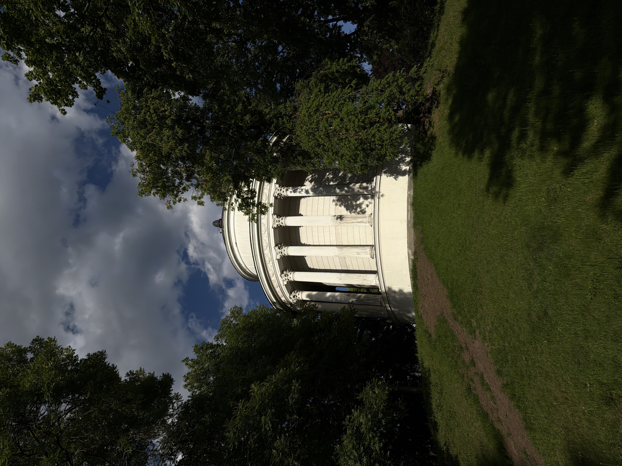 The image shows a rotunda-style building with columns, partially obscured by trees. The building is located on a grassy area with a path. The sky is visible through the trees, with clouds. The location is in the city of Unknown, Unknown.