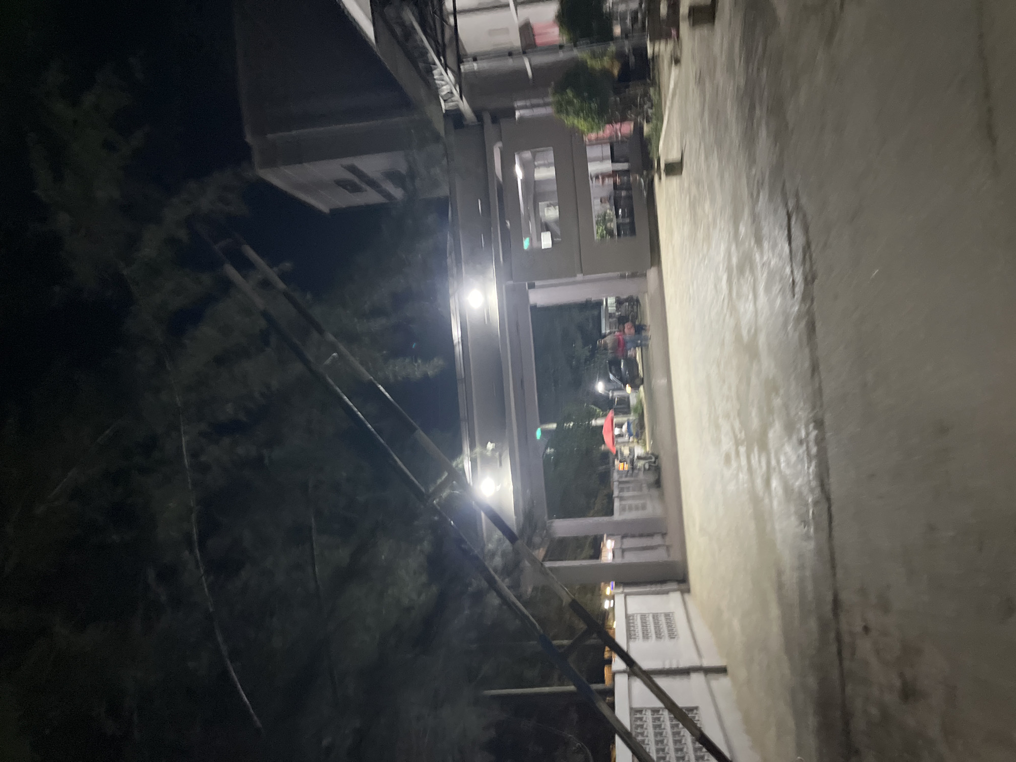The image depicts a dimly lit scene at night, possibly in an urban or semi-urban setting. A wet, reflective pathway or ground dominates the right side of the frame, suggesting recent rain or a damp surface. To the left, a building with multiple stories is visible, with lit windows indicating occupancy. The building has a modern architectural style with horizontal and vertical elements.
In the background, beyond the pathway and between sections of the building, there are glimpses of outdoor activity. A red umbrella is prominently displayed, and several figures appear to be gathered or walking. The lighting is artificial, with streetlights or building lights casting glows and reflections.
The foreground features what looks like a dark, bushy area with some man-made structures, possibly scaffolding or a railing, running diagonally across the frame. This contrast between the natural elements and the man-made structures adds to the complexity of the scene. The overall mood is somewhat mysterious due to the low light conditions and the blurriness of some elements, creating a sense of depth and intrigue. The time of day is clearly nighttime. No text is visible in the image. The context provided of "Kesehatanbandar, Indonesia" suggests a possible location, but specific landmark identifiers are not present in the visual information.