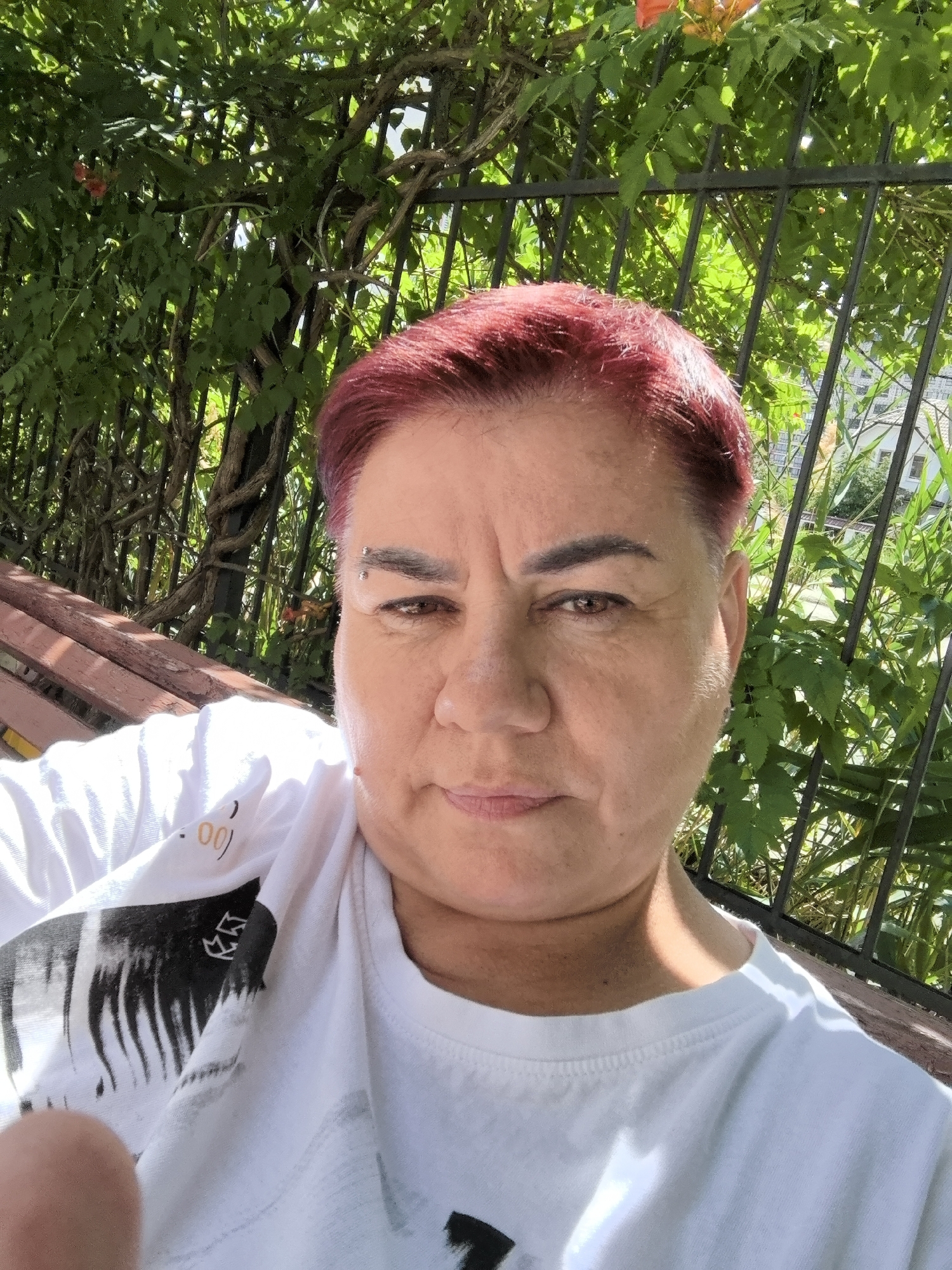 A medium shot shows a person with short, reddish-pink hair and a slight smile, sitting outdoors on a park bench. They are wearing a white t-shirt with a black graphic print, possibly of palm trees. The background is lush green foliage intertwined with a dark metal fence, suggesting a park or garden setting. Sunlight filters through the leaves, indicating it is daytime, likely a warm day. The person has a piercing in their left eyebrow. A hint of red flowers can be seen among the leaves in the upper left corner. The setting appears to be in a moderately urban area, given the fence and hints of buildings in the background to the right. The overall mood is relaxed and casual. The image is a selfie, with the person looking directly at the camera. No text is clearly visible on the person's clothing, although there are some blurred markings on the t-shirt.