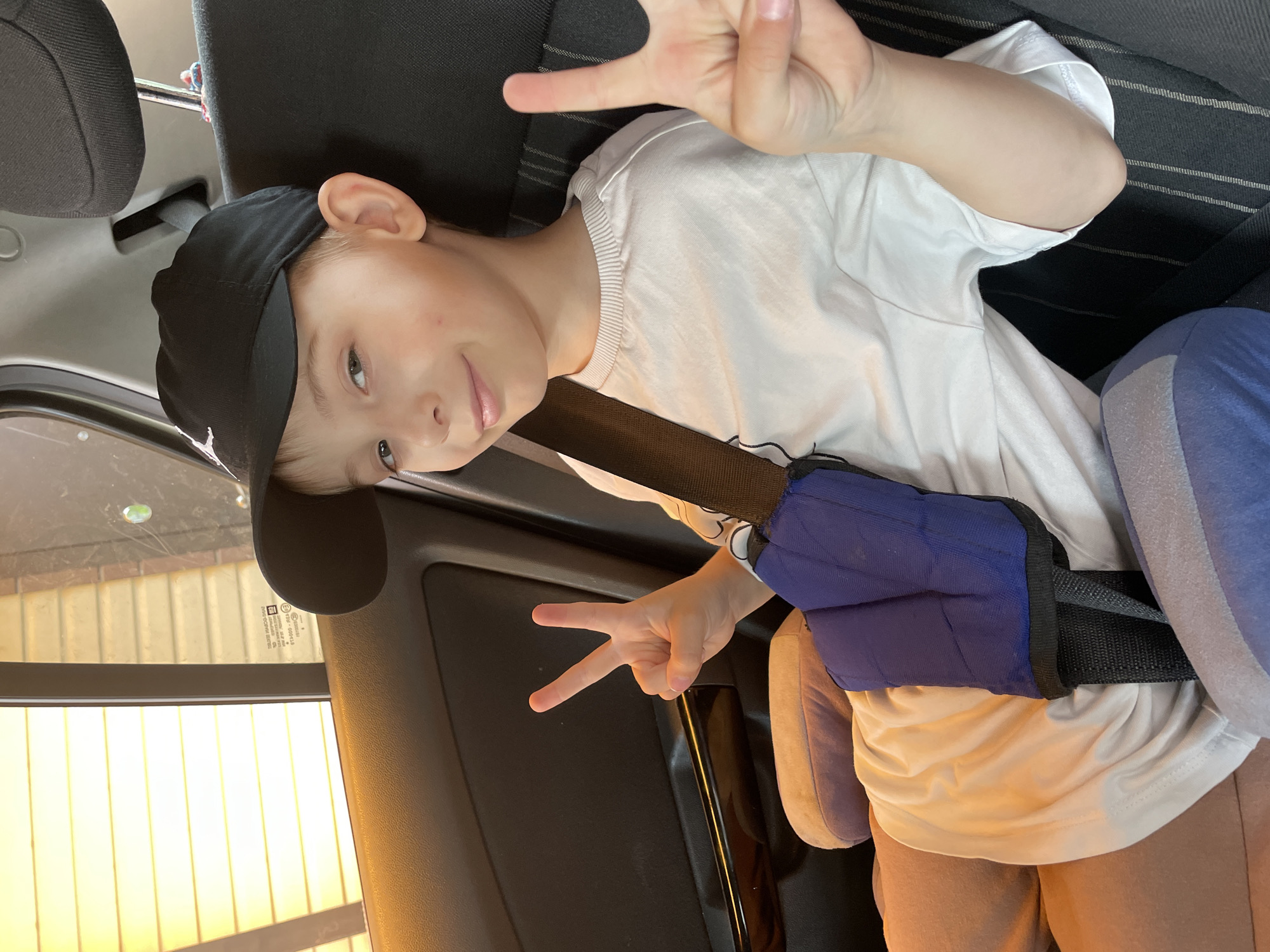 A boy is sitting in a car. He wears a black baseball cap, a white t-shirt, and sweatpants. He is making a "peace" sign with both of his hands. He is wearing a seatbelt with a blue and black cushion on the shoulder strap. The car is parked, and there is a view through the window of a building with vertical slats. The location is unknown.