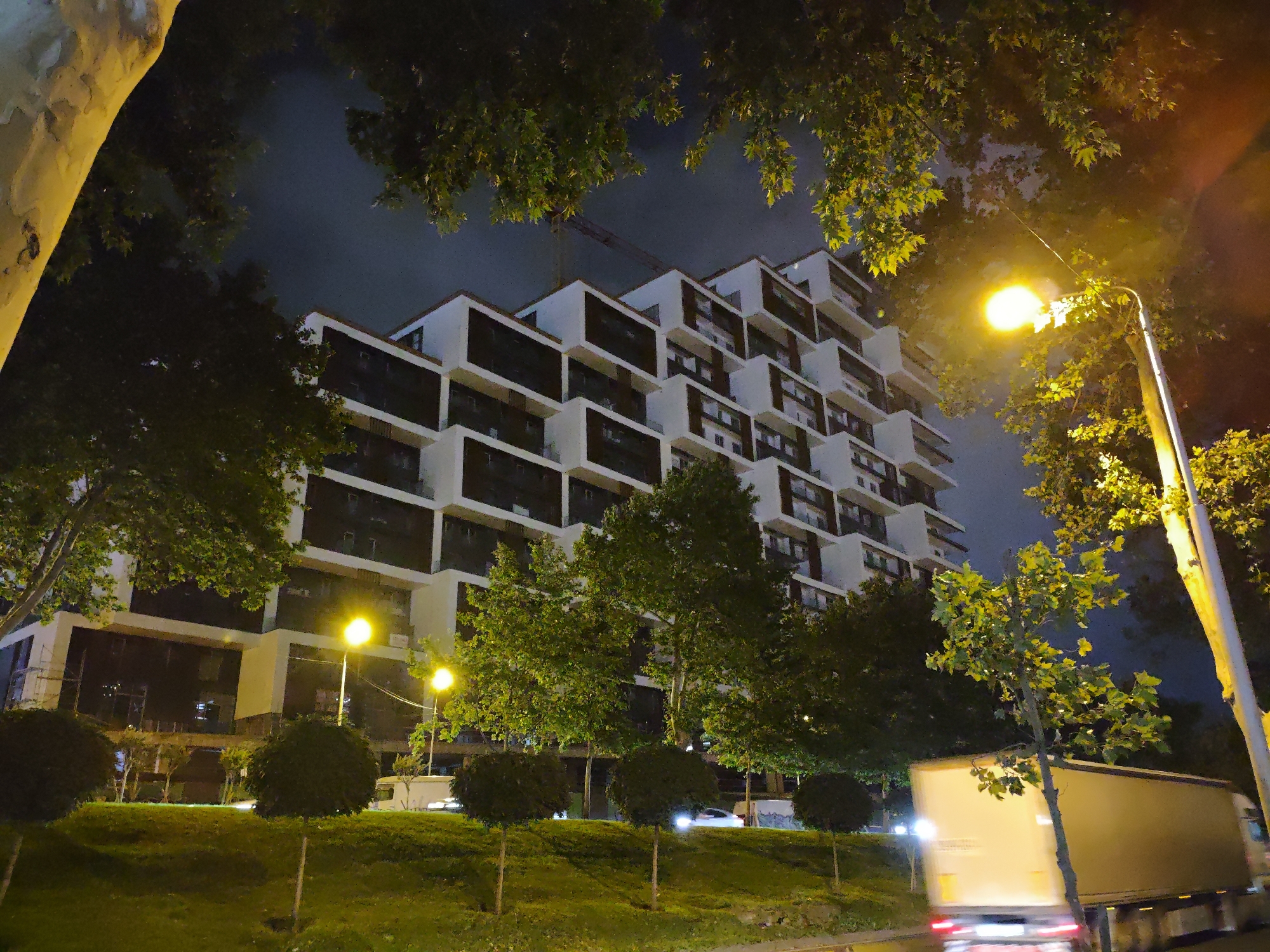 A nighttime exterior shot captures a modern apartment building with a stepped facade and dark windows. In the foreground, a grassy hill is dotted with small, manicured trees, and a large truck is moving along a road to the right. A street lamp illuminates the scene with a warm glow, and trees with lush green leaves frame the top of the image. The sky is dark, with hints of light pollution and a visible construction crane in the distance above the building.