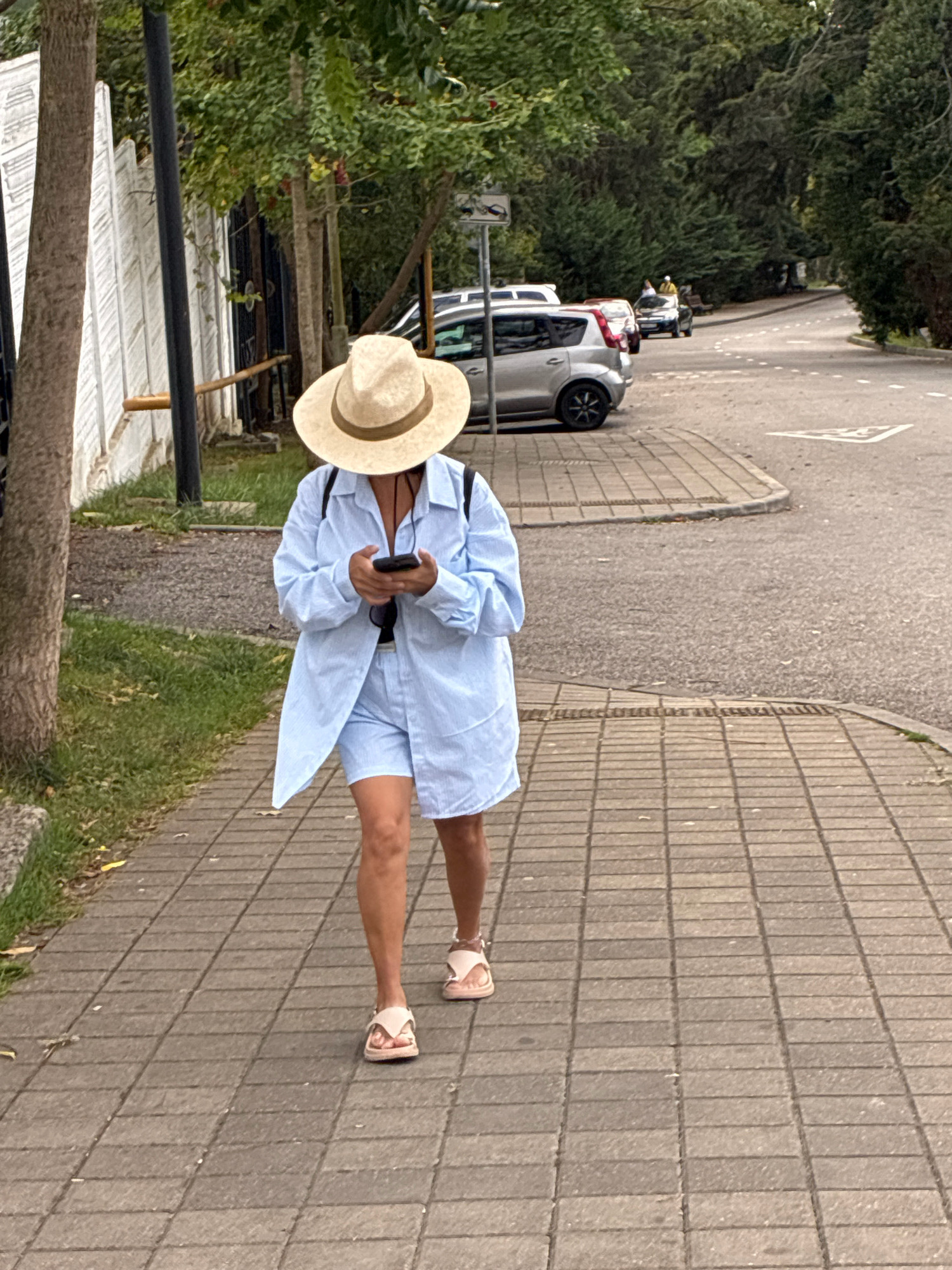 A person wearing a light blue striped button-down shirt and matching shorts, a wide-brimmed straw hat, and light pink sandals walks along a paved sidewalk. The person is looking down at a black smartphone held in their hands. A black backpack is visible on their shoulders. The sidewalk is lined with grass and trees on the left, and a white fence with dark metal gates. In the background, several cars are parked on the right side of a road, which curves away into a densely wooded area. The scene appears to be a park or residential street in Sochi, Russia.