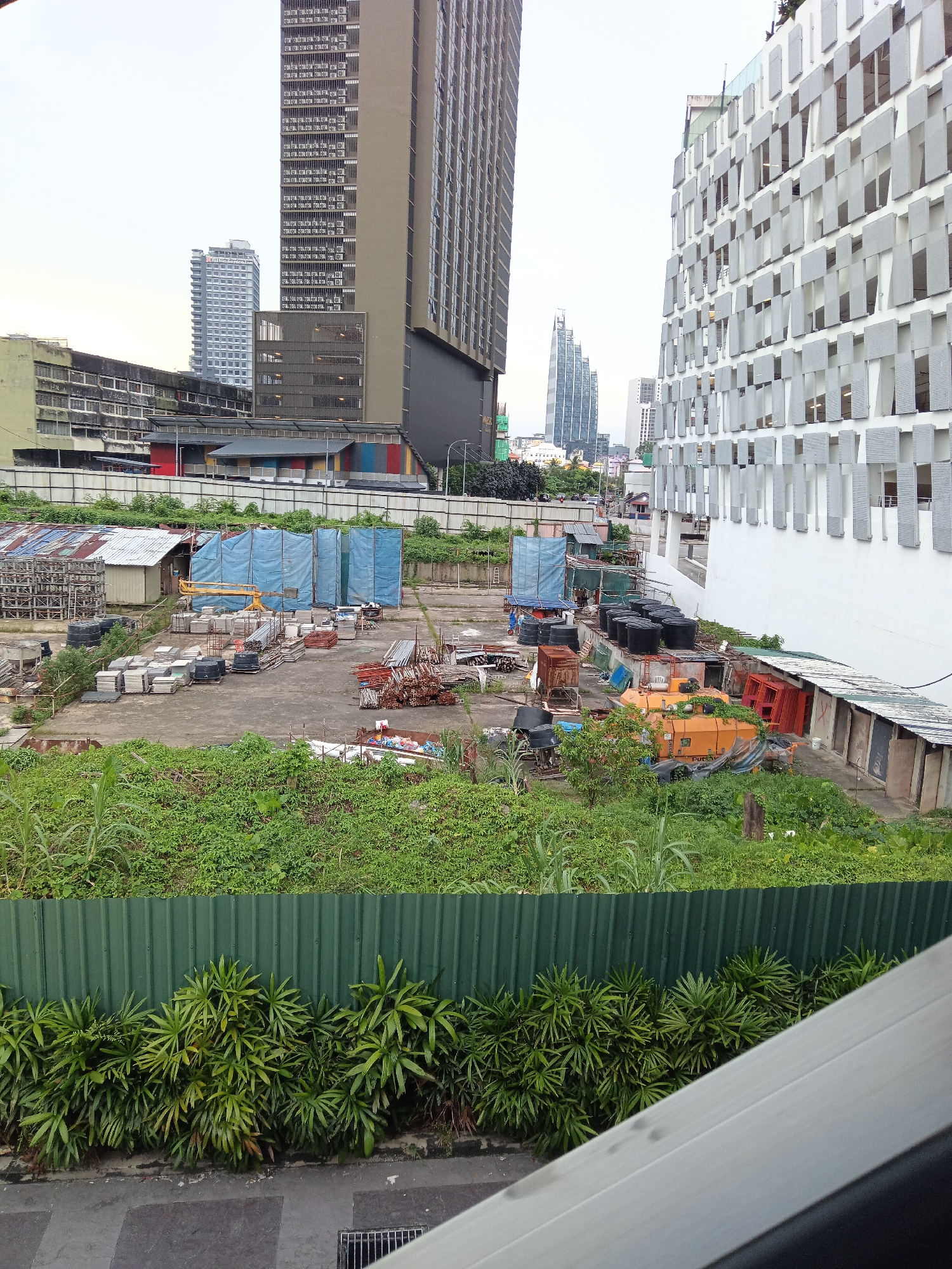 The image depicts an outdoor construction site or storage yard within the city of Kuala Lumpur, Malaysia. In the background, towering modern buildings, characteristic of a metropolitan skyline, are visible. To the right, a distinct white building with a facade of numerous vertical grey panels dominates a section of the frame. In the mid-ground, the area is filled with various construction materials and equipment. This includes stacks of concrete slabs, pipes, rebar, construction barriers, and tarpaulin-covered structures, suggesting ongoing or paused construction work. An orange construction vehicle, possibly a concrete mixer, is also present. Lush green vegetation, including tall grass and shrubs, grows in the foreground and surrounds the construction site, separated by a corrugated green metal fence. The overall scene conveys a sense of urban development interspersed with natural overgrowth.