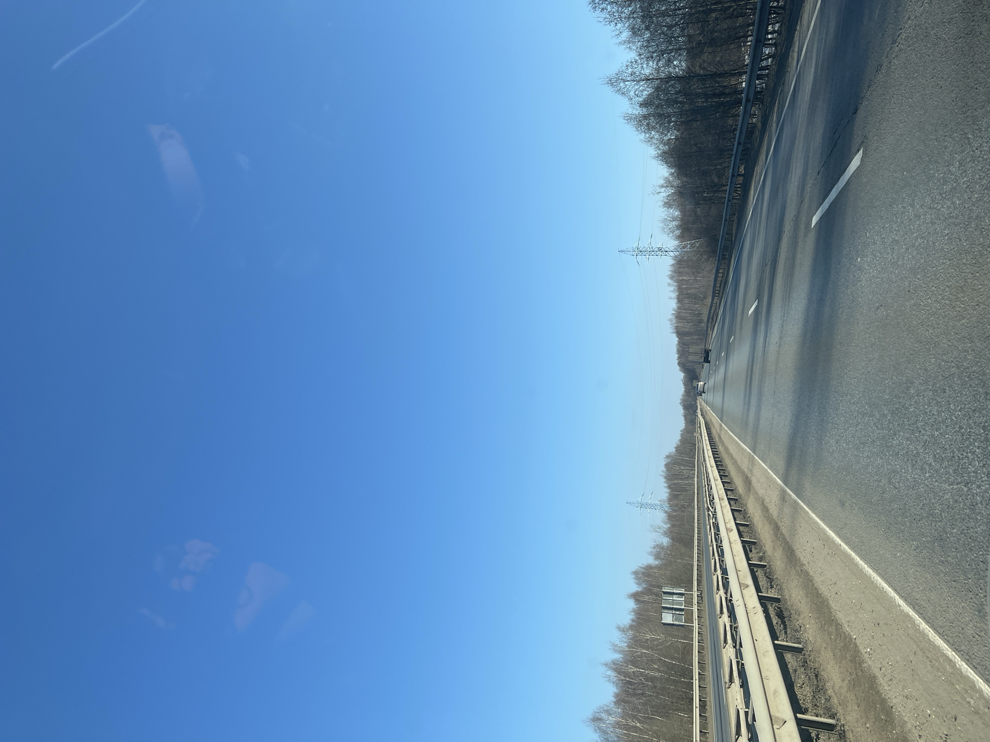 The image depicts a multi-lane highway in Родничок-1 Снт, Russia, under a clear blue sky. The viewpoint is from inside a vehicle traveling on the highway, evidenced by indistinct reflections visible in the upper portion of the sky.
The highway consists of at least two lanes of asphalt pavement visible in the direction of travel, marked by solid white lines on the right shoulder and dashed white lane dividers. A metal guardrail runs along the right side of the road, bordering a deciduous forest of bare trees. A concrete median barrier, topped with an additional metal guardrail, separates the visible lanes from opposing traffic lanes (partially visible) on the left. The base of the median barrier shows accumulated dirt or debris.
Beyond the median barrier to the left, and further beyond the right guardrail, are dense deciduous forests with mostly bare trees, indicating late winter or early spring conditions. Several large, lattice-structured electricity pylons are visible against the sky above the tree line on both sides of the highway in the distance. A dark, rectangular sign structure is mounted above the median barrier in the mid-ground on the left, with unreadable content.
Multiple vehicles are visible as dark shapes in the distance, traveling in the same direction on the highway. Long shadows cast across the roadway suggest a low sun angle, consistent with morning or late afternoon. No individuals are explicitly discernible in the scene.