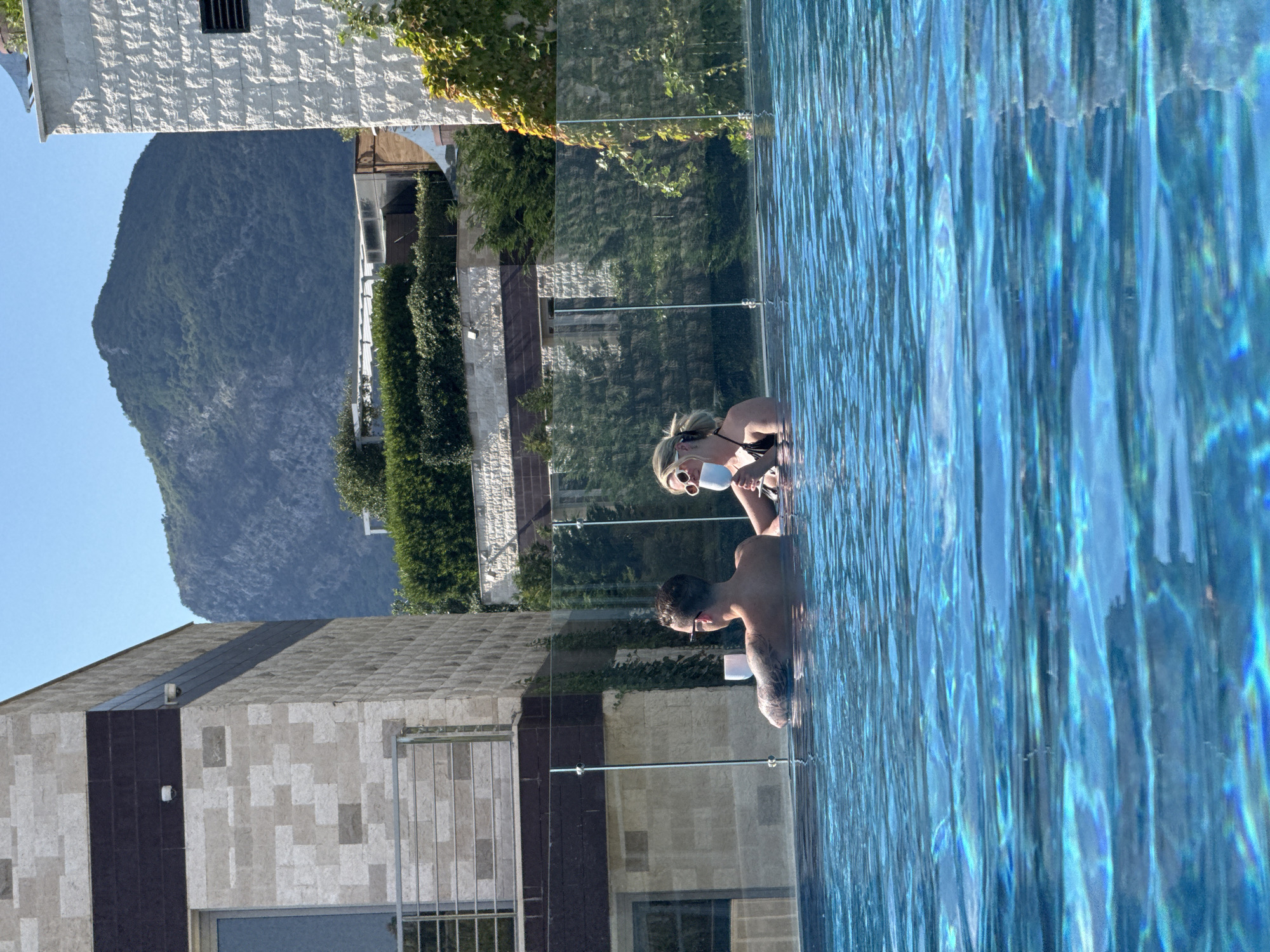Two individuals, a man and a woman, are submerged in a swimming pool, holding white cups. The man, on the left, has tattoos on his arm and wears glasses. The woman, on the right, has blonde hair and wears a black bikini. They appear to be in a resort setting, with modern architecture and lush greenery visible in the background. A majestic, tree-covered mountain looms in the distance, characteristic of Budva, Montenegro. The water's surface is rippling, reflecting the bright sunlight and creating a shimmering effect. The scene suggests a leisurely and relaxed atmosphere in a picturesque location.