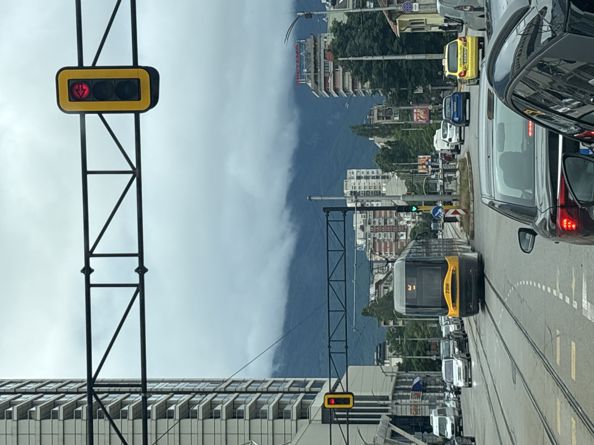 This is a street-level, eye-level photograph taken on a cloudy day in Sofia, Bulgaria. The scene depicts a busy intersection with traffic, a tram, and urban infrastructure. In the foreground, the rear of a black car is visible, with another black car in front of it. Several other vehicles, including cars and a white van, are stopped in traffic. A yellow taxi is also present. The prominent subject in the mid-ground is a yellow and black tram, identified by the number "2356" on its front. The tram is on its tracks, indicating it is either stopped or moving slowly.
The background features a tall, modern, light-colored building on the left, typical of urban architecture. Further back, a range of mountains is visible, partially obscured by thick, low-hanging clouds, suggesting a potential for rain or a generally overcast atmosphere. Across the street, several other buildings of varying architectural styles and heights are present. One building has a red sign that reads "SPETEMA". Traffic lights are visible, with one showing a red light with an arrow pointing left, and another displaying a red light. Overhead, black metal structures, likely part of an electrical system for the tram or traffic control, span across the road. The overall impression is of a typical urban environment experiencing moderate traffic under a heavy, cloudy sky, with a mountain backdrop.