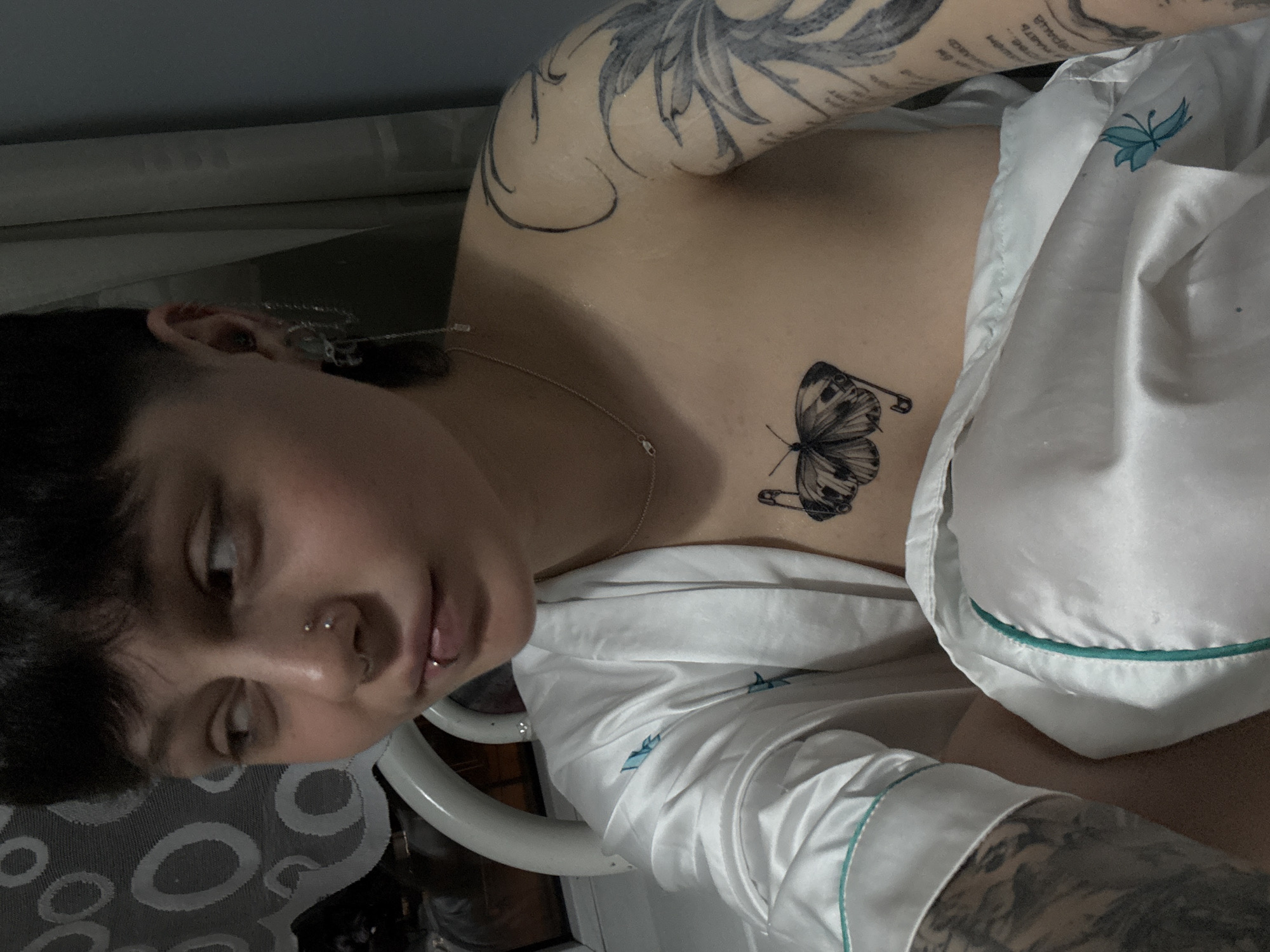A person is lying in bed, facing the camera. They have short dark hair and multiple piercings in their nose and lip. Tattoos are visible on their upper arms and chest, including a butterfly design on the chest. The person is wearing white pajamas with blue trim. The location cannot be determined from the image. There is no evidence to suggest the location is Saint Petersburg, Russia.