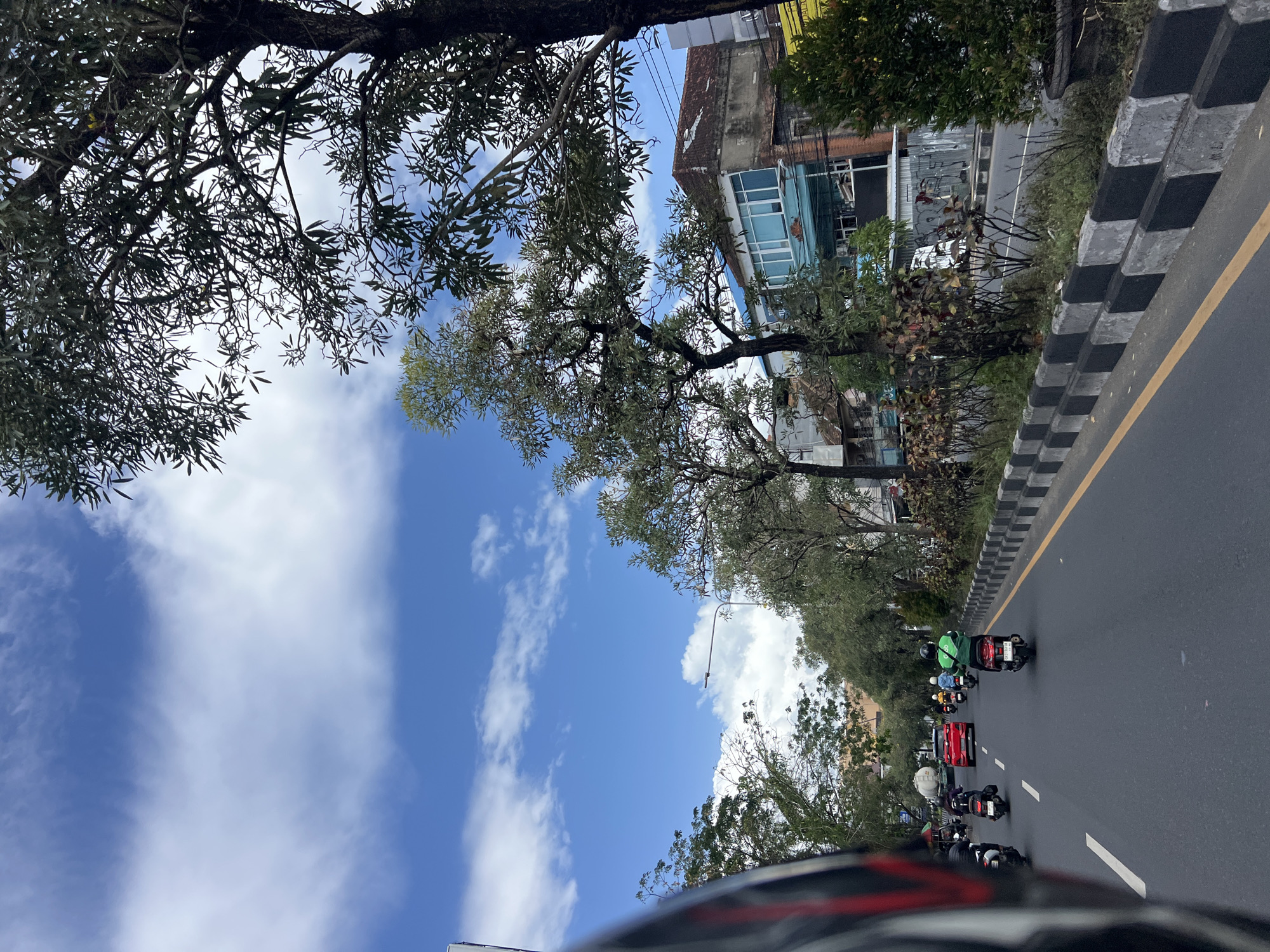 The image captures a street scene in Denpasar, Indonesia, viewed from the perspective of someone riding a motorcycle. The road is paved and marked with white lane lines and a yellow divider. Several motorcycles and a red car are visible on the road, suggesting moderate traffic. The rider's helmet is partially in the foreground on the left. Along the right side of the road, there is a concrete barrier with alternating black and white painted sections, followed by vegetation and buildings. Trees with lush green leaves line the street, providing shade. The sky is blue with scattered white clouds. The architecture of the buildings appears to be a mix of older and more modern styles.