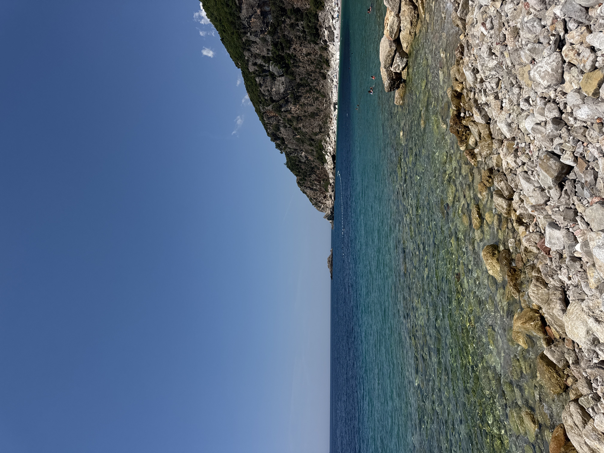 The image depicts a view of the coastline at Perazića Do, Montenegro. The scene is dominated by a clear, blue sky and the turquoise waters of the Adriatic Sea. The foreground is a rocky beach with numerous grey and beige stones of various sizes. Some larger rocks are present near the water's edge.
The water is remarkably clear, allowing visibility of the submerged rocky seabed. In the middle ground, several people are swimming and wading in the sea. Further out, the water transitions to a deeper, darker blue.
A prominent feature of the background is a steep, green-covered mountain slope that curves along the coastline. The mountain is textured with rock faces and patches of vegetation. A small, rocky islet is visible in the distance, further out in the sea, with a line of buoys or markers extending from the shore towards it. In the far distance, the coastline appears to extend further, with a small beach area populated by numerous sunbathers. A few wispy clouds are present in the upper right corner of the sky.