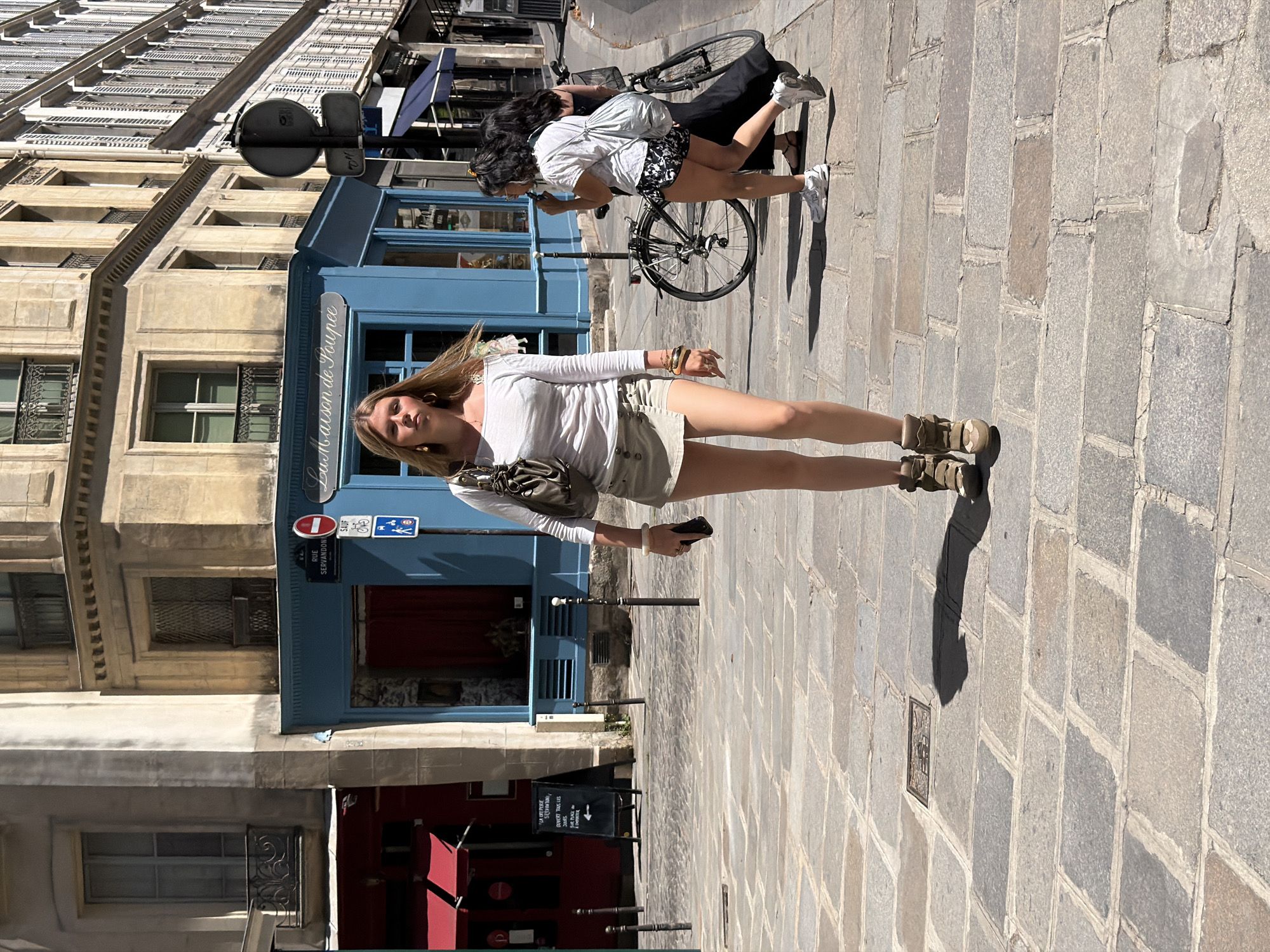 The image depicts a street scene in Paris, France, likely in a commercial area with traditional Parisian architecture. A young woman with blonde hair, wearing a white off-the-shoulder top, a light-colored mini-skirt, and tan wedge boots, walks towards the viewer. She carries a dark green shoulder bag and has bracelets on her left wrist. In the background, a blue shop front with the text "La Maison de Poupée" is visible. To the right of the woman, another woman with dark hair, wearing a white t-shirt and patterned shorts, walks away from the camera with a bicycle. The street is paved with cobblestones, and there are various shops and buildings lining the street, with balconies and shutters on the facades. The lighting suggests a sunny day.
