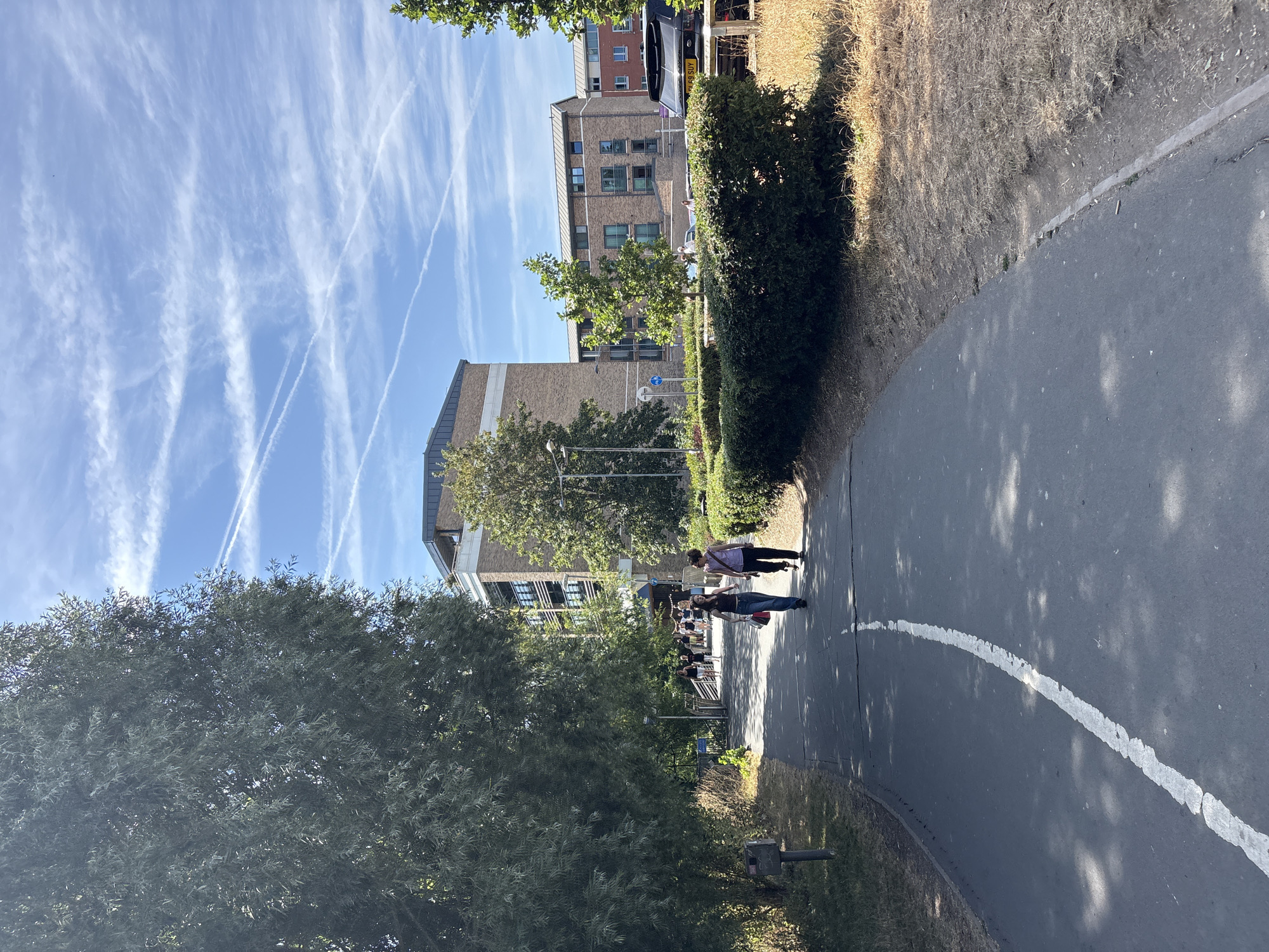 This image depicts a paved walkway in Chelmsford, United Kingdom, bordered by dry grass and a meticulously maintained hedge on the right, and a large tree on the left. In the background, several multi-story brick buildings are visible, some with modern architectural elements like balconies and large windows. The sky is a clear blue, crisscrossed with numerous white contrails, suggesting air traffic. Two individuals are walking away from the camera on the walkway, one carrying a bag. Further down the path, more people are visible near the entrance to the buildings. A black SUV is parked on the right side of the frame, near the buildings. The overall scene appears to be an urban or suburban setting on a sunny day.