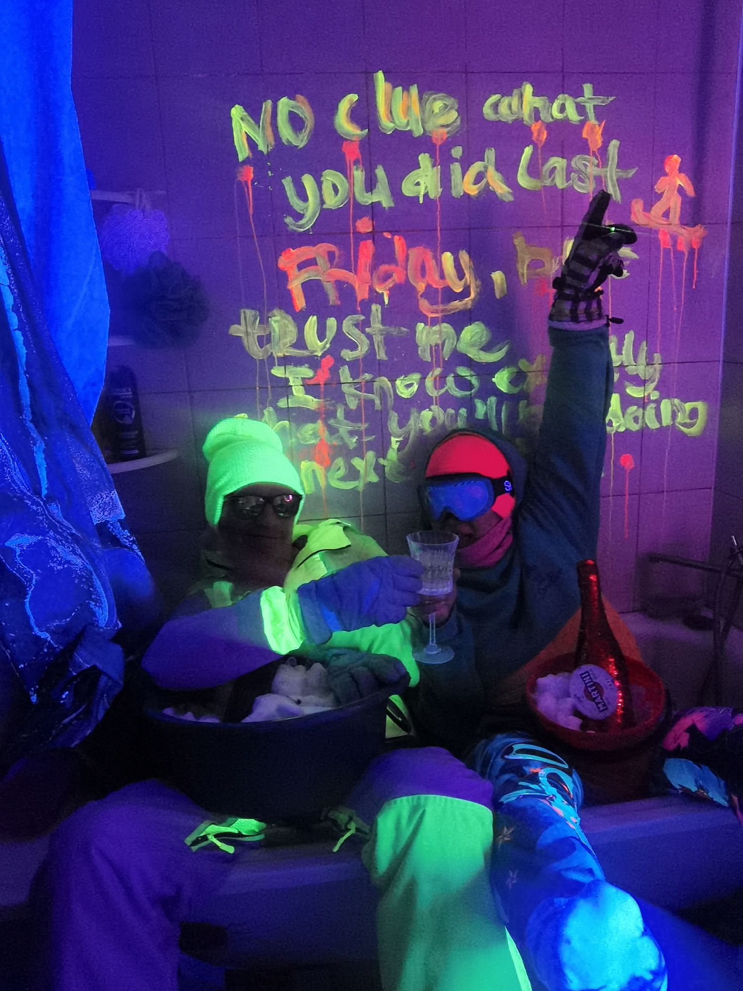 Here is a detailed description of the image:
**General Overview:**
The image depicts two individuals in a brightly lit, indoor scene under blacklight, possibly a bathroom. Graffiti-like writing is visible on the tiled wall behind them. The overall atmosphere is celebratory and somewhat chaotic.
**People:**
* Two individuals are in the foreground.
* One is wearing a neon green beanie, glasses, and a neon yellow jacket. This person is holding a large bowl filled with white material, possibly cotton or packing peanuts.
* The other individual wears pink goggles, a turquoise hooded jacket, and striped gloves. One arm is raised in the air holding a peace sign, and the other holds a champagne glass.
**Objects:**
* **Wall:** Tiled wall with neon writing that reads "No clue what you did last Friday, trust me I know what you’ll be doing next." The text appears to be painted or written with neon/UV-reactive paint.
* **Bowl:** A large bowl filled with white filler.
* **Drinkware:** Champagne glass held by the second person.
* **Bottle:** A Martini bottle is nestled in a red bucket also filled with the white filler.
* **Clothing:** The subjects are wearing brightly colored, casual clothing that is likely chosen for its appearance under blacklight. There is a mixture of neon and vibrant colors, including green, pink, and turquoise.
* **Shower Curtain:** A blue patterned shower curtain is partially visible.
* **Accessories:** Gloves, beanie, goggles.
* **Toiletries:** A bottle is placed on a shelf in the background.
* **Toys:** Two bright green plastic lizard toys sit in front of the bowl.
**Environment:**
* **Location:** It appears to be a bathroom, evidenced by the tiles on the wall, shower curtain, and other bathroom objects.
* **Lighting:** The scene is lit with blacklights, which cause the neon colors to glow brightly.
**Actions/Interactions:**
* The subjects appear to be celebrating or partying. One person is holding up a champagne glass, suggesting a toast. The other person seems to be in possession of a bowl filled with filler.
* The overall impression is one of revelry, possibly after a night out.
**City:**
The name of the city, Chaikovsky, Russia, is not visible in the image.