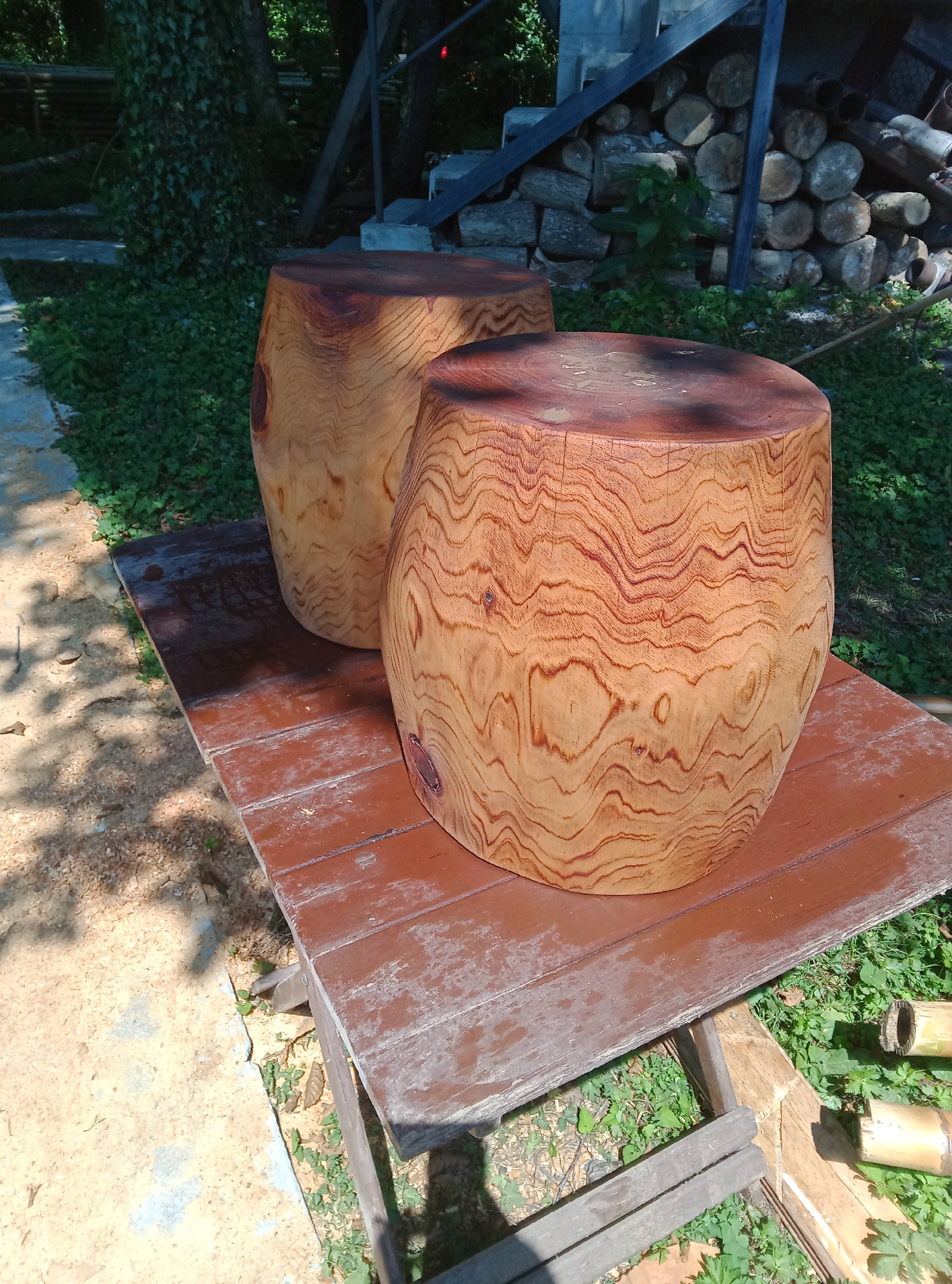 Two large, rough-hewn wooden stumps, likely used as stools or side tables, are placed on a weathered brown wooden platform outdoors. The stumps exhibit distinctive wood grain patterns, with lighter wood interspersed with darker, wavy lines. One stump is closer to the foreground, taking up a significant portion of the frame, while the other is partially visible behind it. The platform itself appears to be made of several wooden planks.
The environment surrounding the platform is natural and slightly unkempt. Green grass and other vegetation grow around and beneath the platform, with some areas of loose, light-colored soil or wood shavings visible to the left. In the background, a stack of cut logs is visible against a structure, and more greenery and trees suggest a wooded or garden setting. The lighting indicates it is daytime, with shadows cast on the platform and the ground. The overall impression is that of an outdoor, rustic setting, possibly a backyard or a workshop area. No people are present in the image. The specific location of Pshap, Abkhazia is not visually discernible from the image content.