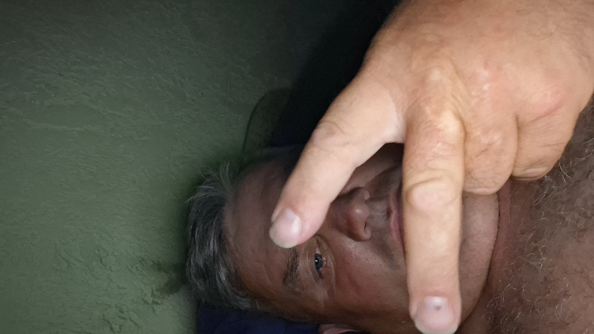 A close-up, low-angle, selfie-style shot shows a man from the chest up, lying down, with his face partially obscured by his right hand which is held up to his face. He has graying hair and is looking at the camera with his left eye visible. His skin appears flushed. The background is a textured, dark green wall. The overall lighting is dim. The image is taken in Voinova, Moldova.