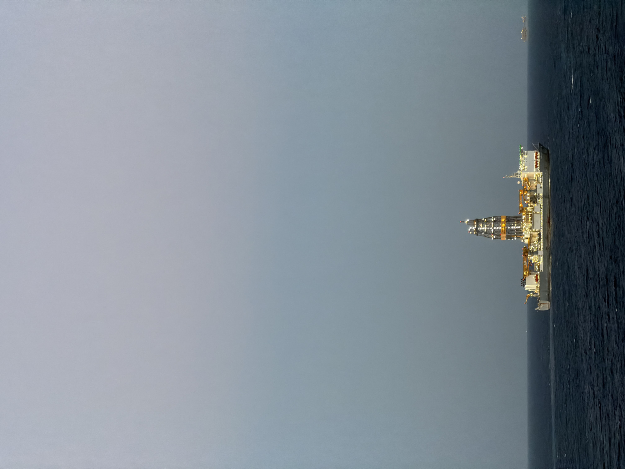 A night time, horizontally oriented, medium shot shows a tanker vessel, lit with yellow lights, floating on a body of water under a grayish sky. The tanker is on the right and the body of water is dark. The majority of the frame is gray. Another ship can be seen in the far distance near the right edge. The exact location is unknown.