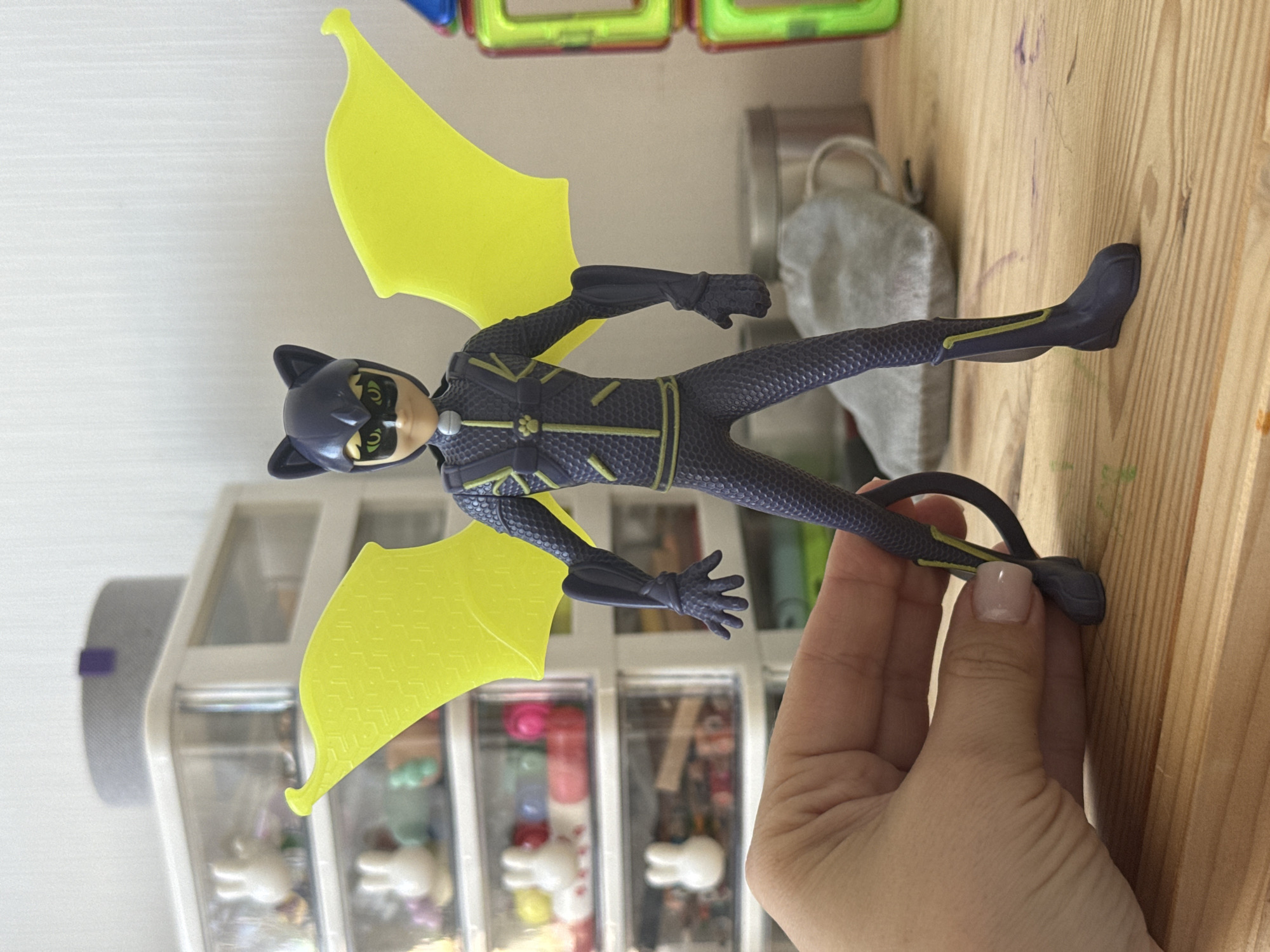 A plastic action figure of a character with a cat mask and yellow bat-like wings is held by a hand with light pink nail polish in the foreground. The figure is dark blue with yellow accents and a textured pattern. In the background, there are clear plastic drawers filled with small objects, a gray cylindrical object, and a stack of colorful magnetic building blocks. The scene is set on a wooden surface. The image was taken in Lyubertsy, Russia.