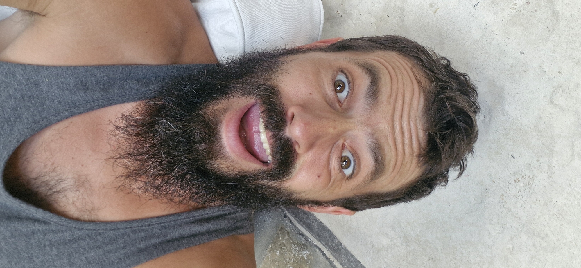 The image shows a close-up, upside-down selfie of a man with a full beard and dark hair. He is wearing a gray tank top. His eyes are wide open, and he has a surprised or excited expression. His mouth is open, showing his teeth and tongue. The man's forehead is heavily wrinkled, suggesting he may be squinting or expressing a strong emotion. The background appears to be a textured, light-colored surface, possibly concrete or stone, with some dark smudges or marks. A small portion of a white object, possibly a pillow or piece of furniture, is visible in the upper left corner. The lighting is bright, suggesting it is daytime. There is no visible text in the image.