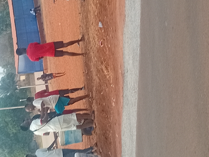 The image depicts an outdoor scene in Ayangba, Nigeria, featuring an unpaved reddish-brown dirt area next to a paved gray road. Seven individuals are present. In the left-foreground, a person in a white t-shirt and white shorts, carrying a dark shoulder bag, stands adjacent to a light-colored cooler on wheels. Immediately to their right, a second individual in a light-colored t-shirt and blue shorts is captured in mid-motion with their left leg raised and bent, facing the first person. To the right of these two, a third person in a red t-shirt and dark shorts stands with their back to the viewer. Further back, a fourth person in a red t-shirt and dark shorts is partially visible. A prominent blue rectangular building with a red top trim stands in the mid-background to the right. A fifth individual in a patterned dark shirt and shorts stands on the dirt in front of the blue building. In the far background, a sixth person in a white shirt and dark pants is seated near the base of the blue building. Two slender light poles are visible in the background to the left of the building. Distant green foliage frames the upper background. The dirt ground contains scattered small white debris. The lower portion of the image shows a gray paved road with a distinct lighter gray edge marking the transition to the dirt area.
