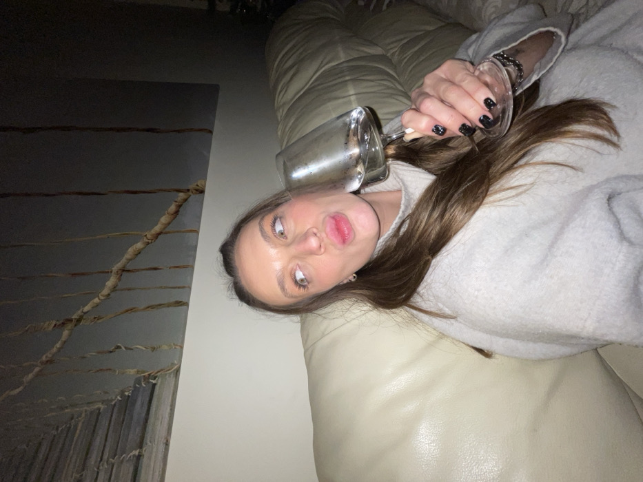 A woman with light brown hair lies on a tan couch. She is wearing a light gray sweater. She holds an upside-down clear glass filled with liquid near her face, which is tilted. Her lips are puckered. She has dark nail polish. In the background, there is a gray wall with a textured pattern resembling a horizontal abstract bridge. The lighting in the room appears dim.