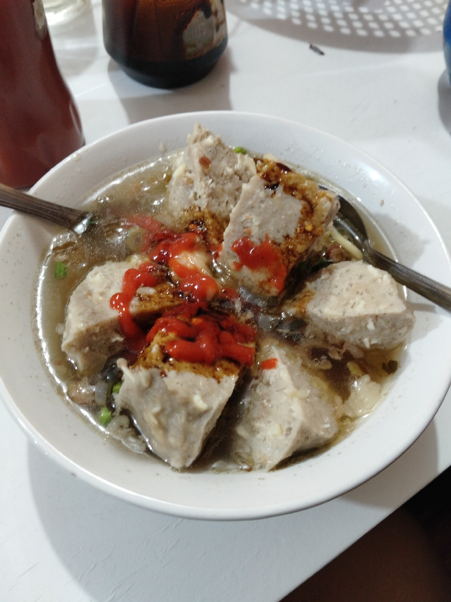 A close-up, eye-level, indoor shot shows a white bowl of bakso, a type of Indonesian meatball soup, on a white surface. The bowl contains several large, round, pale-gray meatballs, sliced in half, submerged in a clear, light-brown broth. A swirl of bright red chili sauce is drizzled over the meatballs, and a darker, soy-sauce-like condiment is also visible. Pieces of green onion are scattered in the broth. A spoon with a dark handle rests on the left side of the bowl, partially submerged, and another spoon is visible on the right side. In the background, out of focus, is a dark brown bottle with a patterned lid and a red bottle with a metallic cap. The overall lighting suggests an indoor setting with artificial light. The image appears to have been taken in the city of Sokaraja, Indonesia, a known culinary hub for bakso.