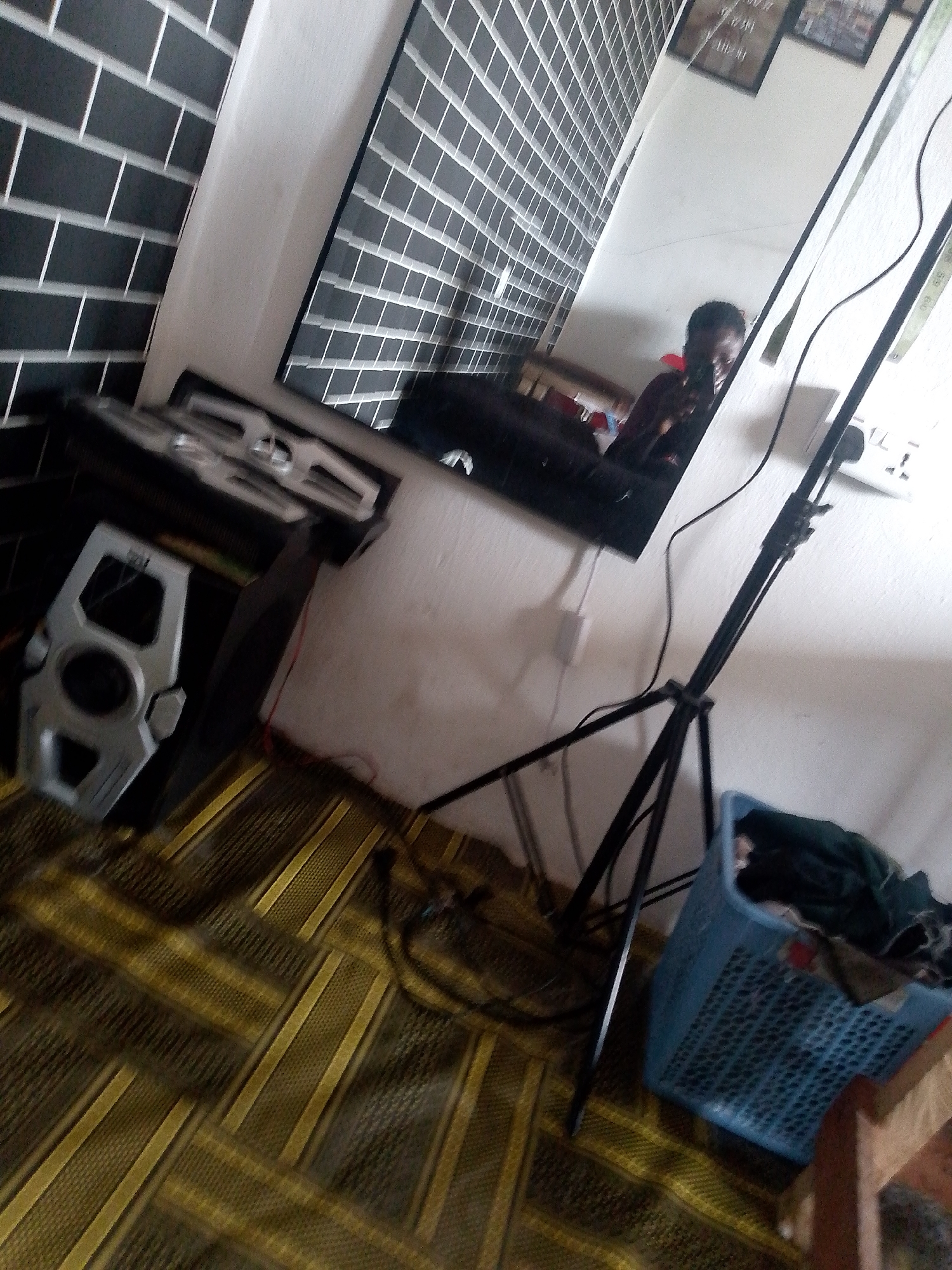 This is a first-person perspective image, likely taken by someone using a phone, in a room in Jauro Yinu, Nigeria. The scene appears to be a domestic setting, possibly a bedroom or living area, with elements suggesting preparation for or the aftermath of an event like a photoshoot or filming.
Dominating the left side of the frame is a black computer tower, its front panel featuring a silver, geometric grill with a central circular speaker. Above this, a silver-colored component, possibly part of a sound system or another computer peripheral, with parallel horizontal slots, is visible. The background wall is covered in a distinctive black and white brick-patterned wallpaper, with a narrow white vertical trim separating it from a plain white wall on the right.
A large mirror is angled on the right side of the image, reflecting a portion of the room and its occupants. In the mirror's reflection, a person, whose face is mostly obscured, is seen holding a dark object, likely a phone or camera, towards the mirror, indicating they are taking the photo. They are wearing a dark-colored garment. The reflection also shows a white wall with two framed pictures hanging on it.
A black tripod, a common piece of equipment for photography or videography, stands prominently in the middle right of the frame, its legs splayed on the floor. Near the base of the tripod is a bright blue plastic laundry basket filled with dark-colored clothing. Electrical cords are visible, one red and one black, running along the floor.
The floor is covered in a carpet with a repeating pattern of alternating dark and yellow diagonal stripes, creating a herringbone-like design. The lighting in the room is somewhat dim, contributing to the slightly grainy quality of the image. The overall impression is of a somewhat cluttered but functional space.