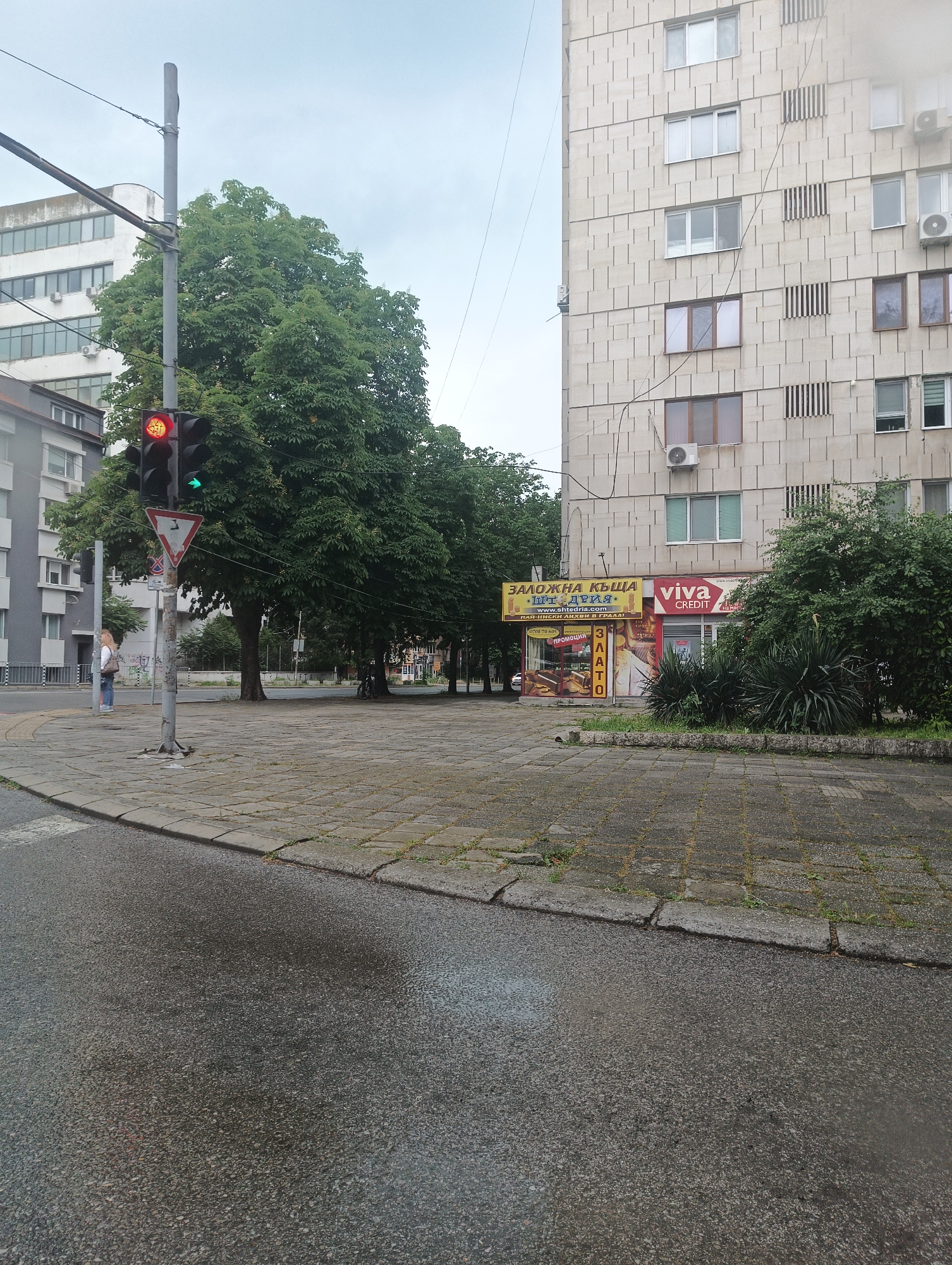 The image depicts a city street scene in Burgas, Bulgaria, notable for the intersection and the buildings lining the street. Dominating the right side of the frame is a tall, light-colored apartment building with a textured facade, featuring both windows and vertical decorative elements. At its base, there are two business signs: one for "ЗАЛОЖНА КЪЩА ШТЕДРИЯ," presumably a pawn shop, and the other for "viva CREDIT."
To the left, a traffic light stands at the corner, currently displaying a red light above a green arrow. A yield sign is also present. A woman is standing on the corner near the traffic light, waiting to cross the street. Trees line the street beyond the intersection, creating a green canopy. The pavement and street appear wet, suggesting recent rain. The street surface is gray and shows signs of wear. In the background, another light-colored building is partially visible behind the trees.