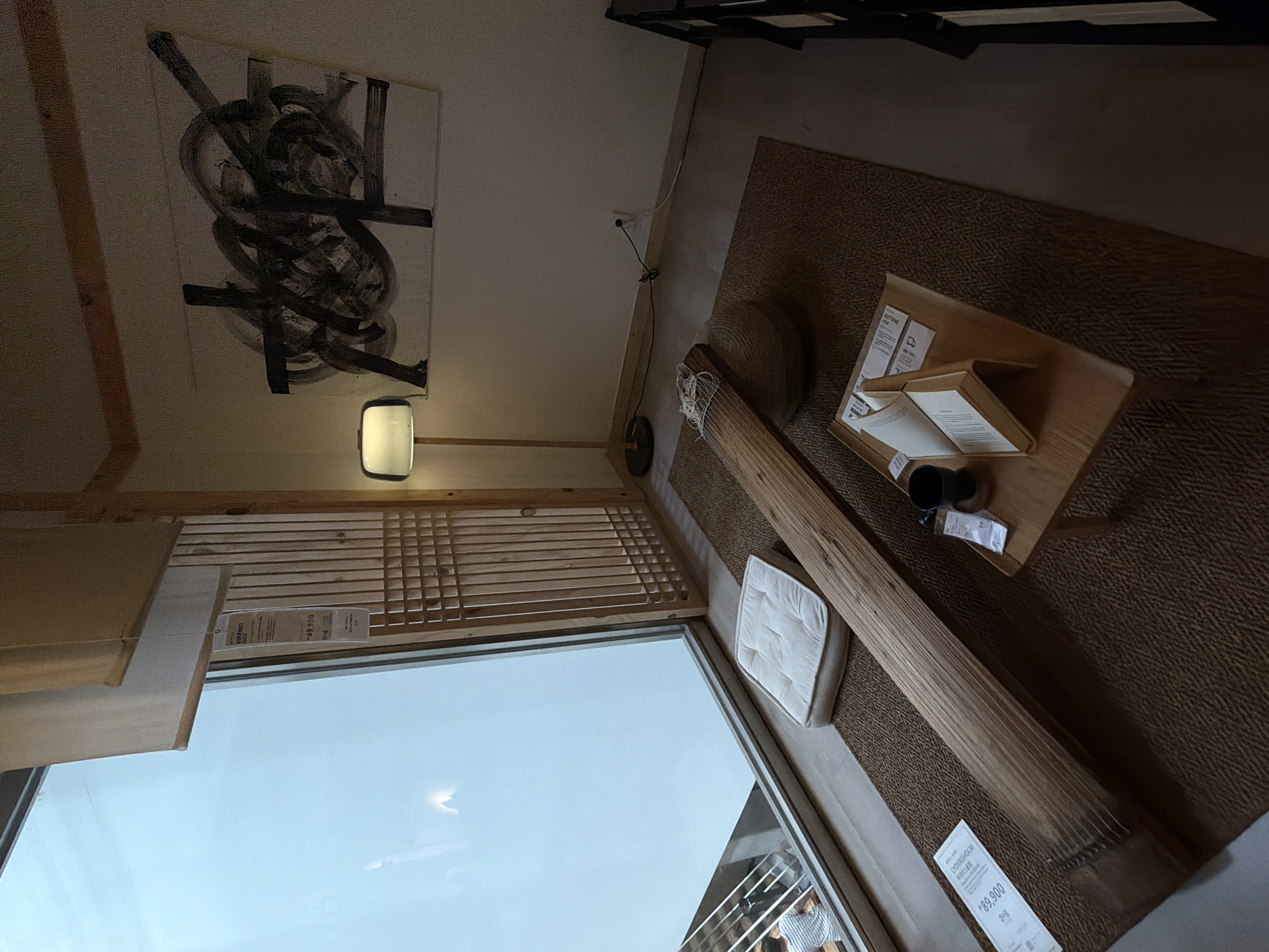 The image depicts an indoor scene, likely within a retail or exhibition space in Goyang-si, South Korea, given the presence of a product tag with Korean text. The room features a large window on the left, with a translucent white covering and wooden interior shutters. A person in a striped shirt is partially visible outside the window.
Dominating the foreground is a large, long, wooden musical instrument, possibly a gayageum, laid horizontally on a textured brown rug. To its left, a white cushioned mat rests on the rug. A small, light-colored wooden table is situated to the right of the instrument, holding an open book, a black mug, and some paperwork.
In the background, a floor lamp with a light-colored rectangular shade stands to the left. On the wall behind the lamp, an abstract painting with black and white brushstrokes is displayed. The wall itself is a neutral color, and an electrical outlet with cords is visible near the baseboard. To the far right, a dark shelving unit is partially visible. The overall lighting is dim, with the exception of the lamp and the light from the window.