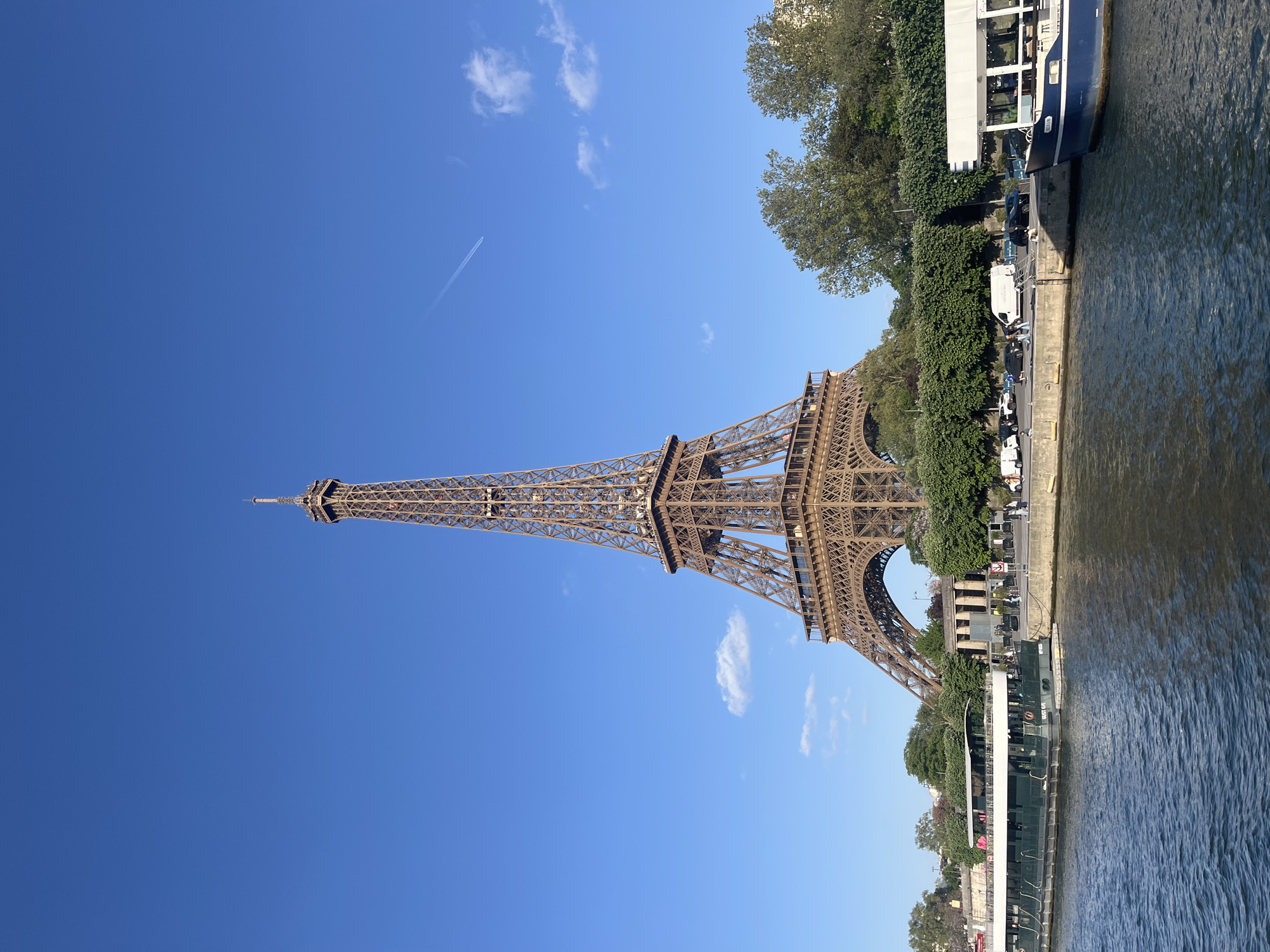 This media file presents a vibrant daytime view of Paris, France, centered around the iconic Eiffel Tower. The colossal iron lattice structure of the Eiffel Tower dominates the left side of the frame, standing tall against a clear, expansive deep blue sky. A few wispy white clouds are scattered across the sky, and a faint white airplane contrail streaks across the upper portion.
To the right of the Eiffel Tower, the calm waters of the Seine River flow, exhibiting subtle ripples and reflections of the bright sunlight. The riverbank is visible between the tower and the river, densely lined with lush green trees and well-maintained shrubs. A road runs along this quay, where several vehicles, including cars and vans, are parked or moving. A number of people can be seen strolling or standing on the riverbank, suggesting pedestrian activity.
Several boats are present on the river. A large, dark blue and white passenger boat, identifiable as a Bateau Mouche, is prominently featured on the right side of the image, either docked or cruising close to the bank. The text "BATEAUX MOUCHES" is faintly visible on its side. Another smaller, possibly green and white, vessel is also seen further along the waterway.
The time of day appears to be late morning or early afternoon, characterized by bright, direct sunlight and a clear weather. The overall scene captures a pleasant and active atmosphere, highlighting the famous Parisian landmark and the bustling life along the Seine River.