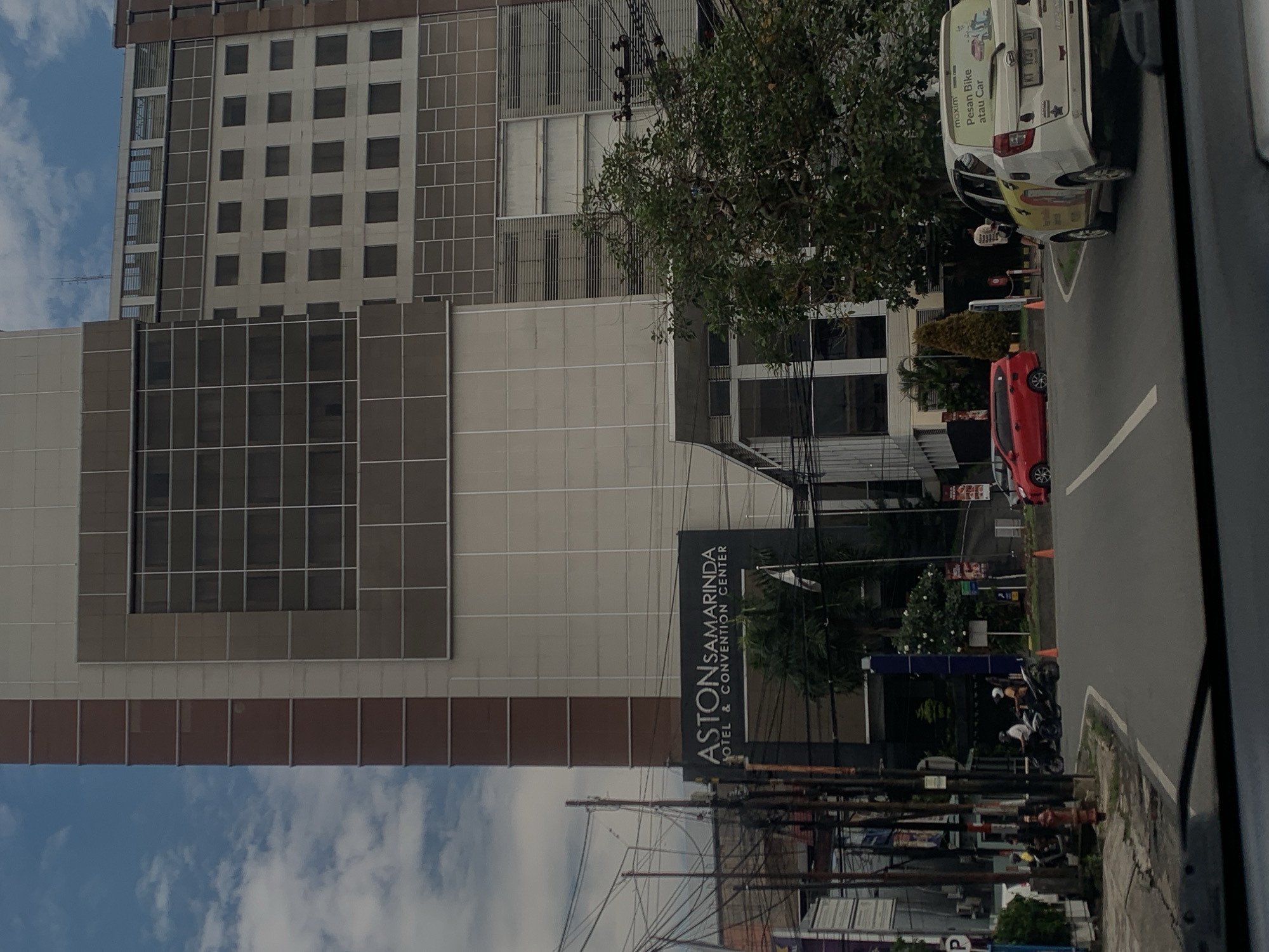 The image shows the Aston Samarinda Hotel & Convention Center in Samarinda, Indonesia. A tall building with a grid-like facade is visible. A sign for the hotel is displayed in front of the building. In the foreground, power lines are visible. On the street, there are vehicles, including cars and motorcycles.
