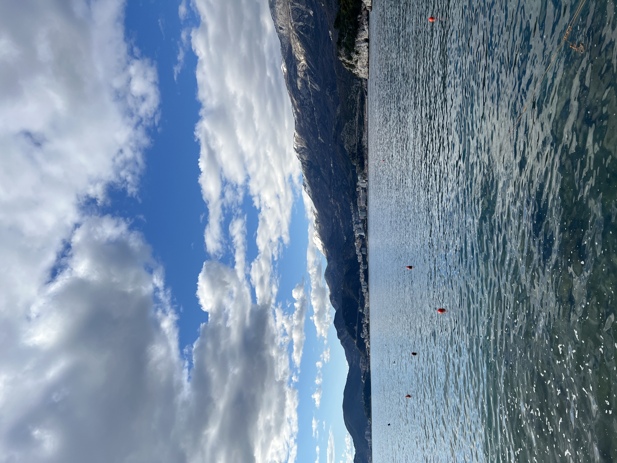 The image shows a landscape featuring a body of water, a mountain range, and a sky with clouds. The water occupies the right side of the image, reflecting light. There are several red buoys visible in the water. The mountain range runs vertically, separating the water from the sky. The sky, on the left side of the image, shows a blue expanse interspersed with various types of clouds, including cumulus and stratocumulus formations. The mountain range is located in the city of Boreti, Montenegro. The image is taken during daylight.