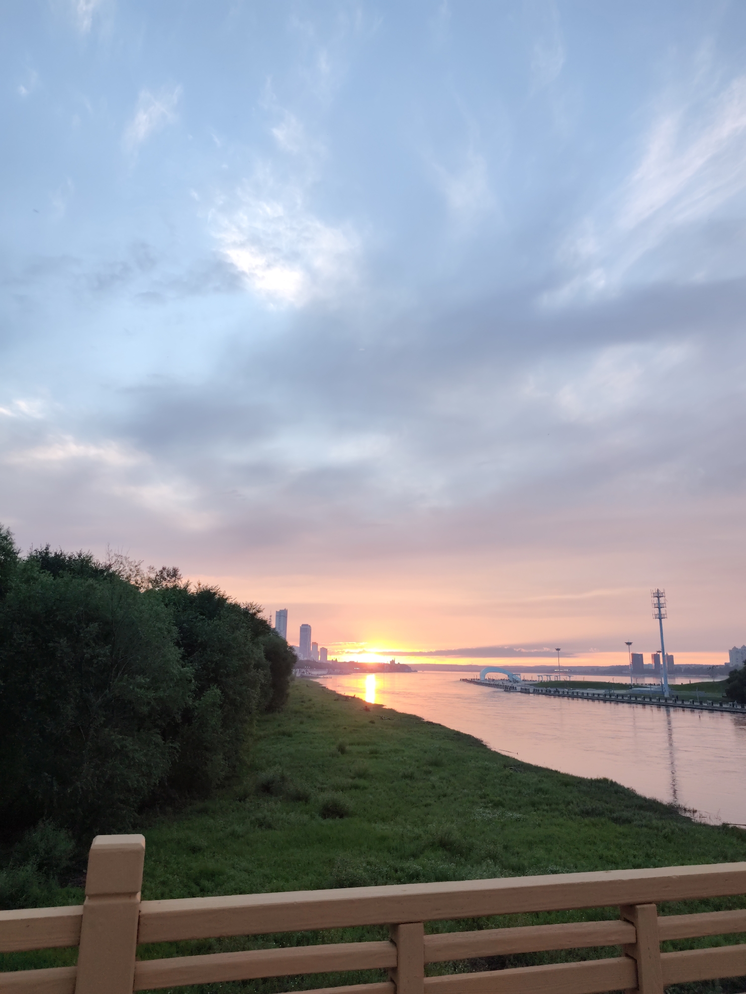 This image depicts a waterfront scene at dusk in 幸福乡, China. A calm river, reflecting the soft hues of the sunset, occupies the right side of the frame. On the left bank, a dense line of dark green trees forms a natural border, transitioning to a grassy embankment in the foreground. In the distance, across the river, a cityscape with tall buildings is visible, silhouetted against the colorful sky. The sky itself is a gradient of soft blues and pinks, with scattered clouds. A wooden railing is visible in the immediate foreground, suggesting the viewpoint is from a raised platform or walkway. There are no people or specific actions captured in this static landscape view.