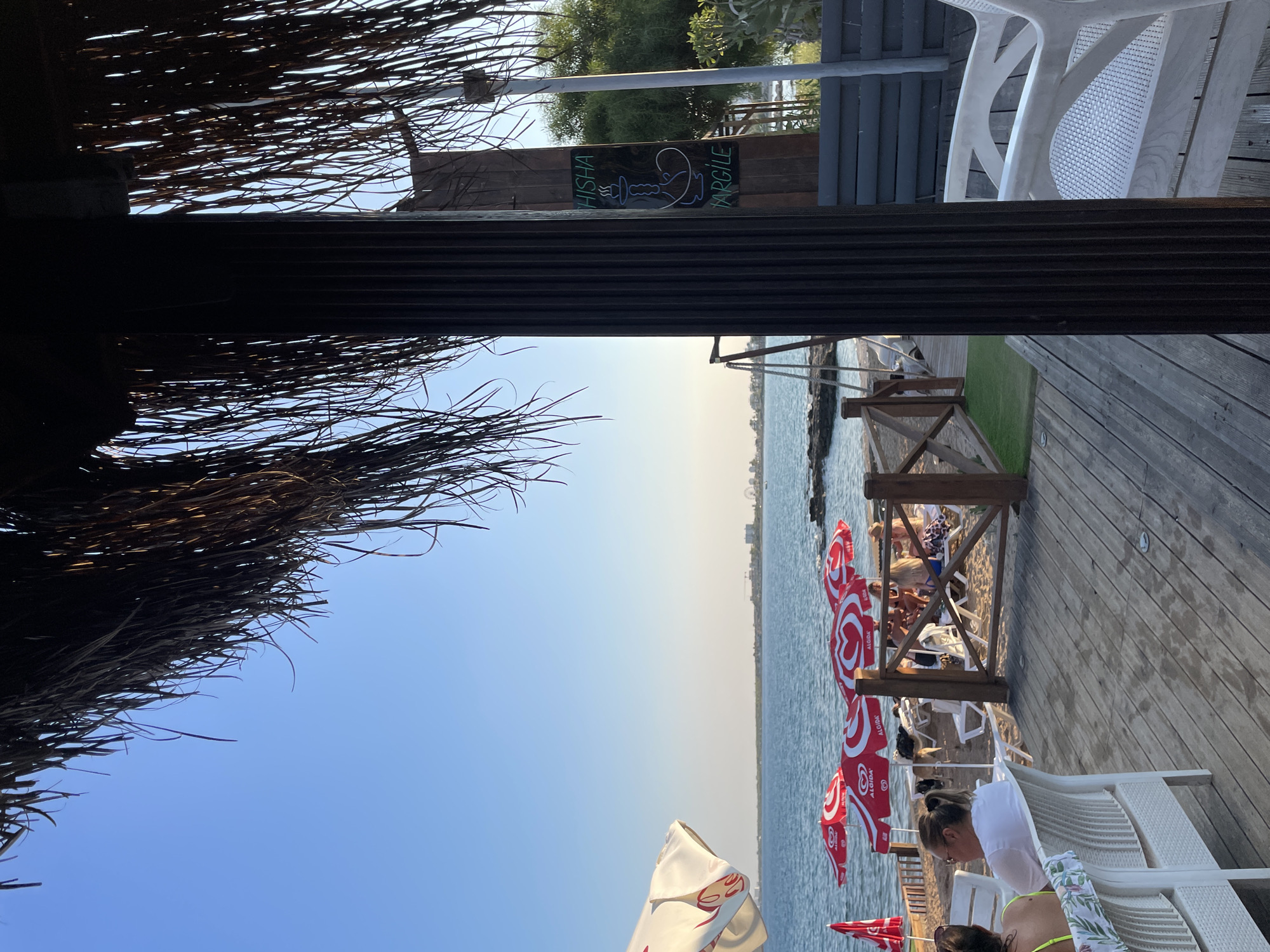 The image depicts a seaside view from a wooden deck, likely a cafe or restaurant, in Trikomo, Northern Cyprus. A thatched roof structure partially obscures the upper part of the frame. The foreground features white outdoor seating and a wooden deck. Beyond the deck, a sandy beach is visible with several red and white beach umbrellas, some bearing the "Algida" logo. Beach chairs are set up on the sand, and a few people are seated on them. The sea stretches out to the horizon, with a distant coastline and buildings visible. To the right, a wooden post with a sign that reads "HISHА TARGILE" with a shisha pipe graphic is present. The overall atmosphere suggests a relaxed, late afternoon or early evening setting by the sea.