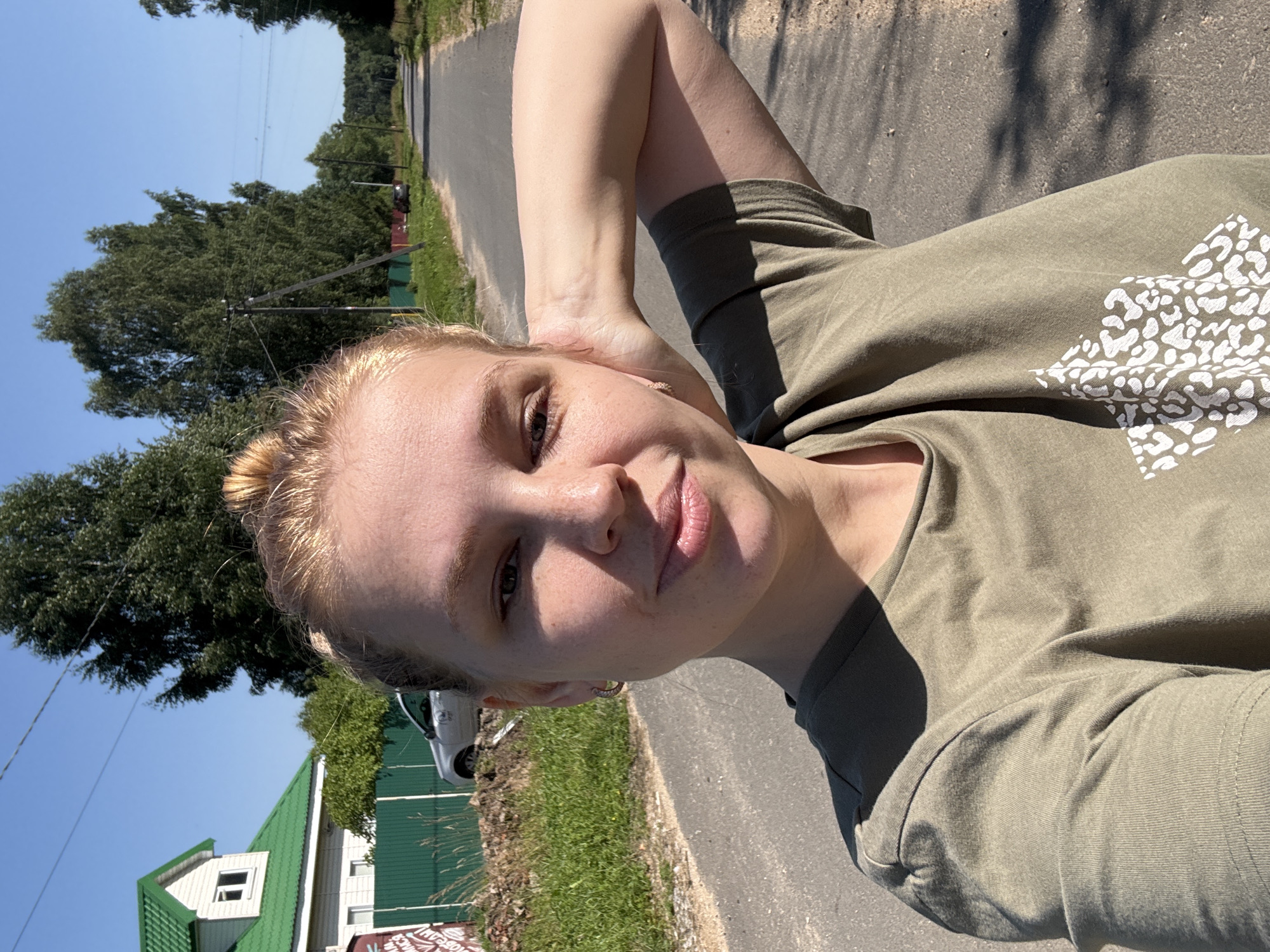 A young woman with blonde hair tied in a bun is taking a selfie on a sunny day in a rural area. She is wearing a green t-shirt with a white pattern on it. Her left arm is bent with her hand resting on the back of her head. The background shows a paved road, a green fence, a house with a green roof, trees, and utility poles. The location is identified as Dunilovo, Russia.