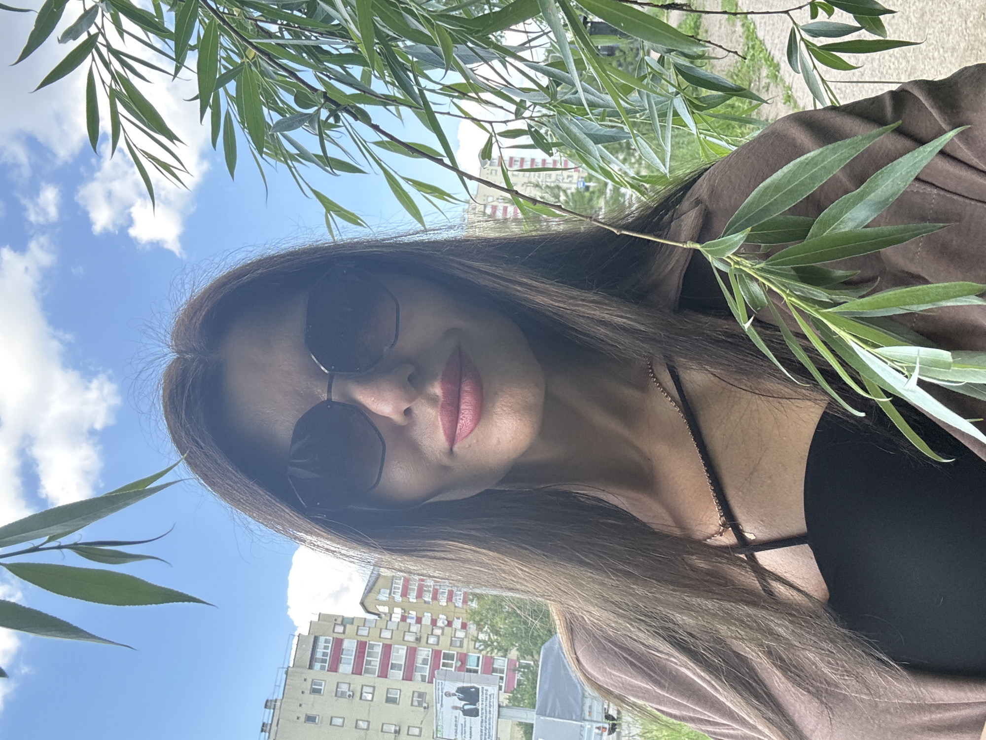 An outdoor, medium shot selfie depicts a woman with long, dark brown hair, wearing dark sunglasses, a brown open jacket, and a black strappy top. Her lips are painted pink. She is positioned in the foreground, with a background that includes a blue sky with scattered clouds, green foliage from a tree obscuring parts of the frame, and residential buildings in the city of Chaikovsky, Russia. A billboard with images of two men in suits is visible on one of the buildings. The overall impression is a candid portrait taken on a bright day.