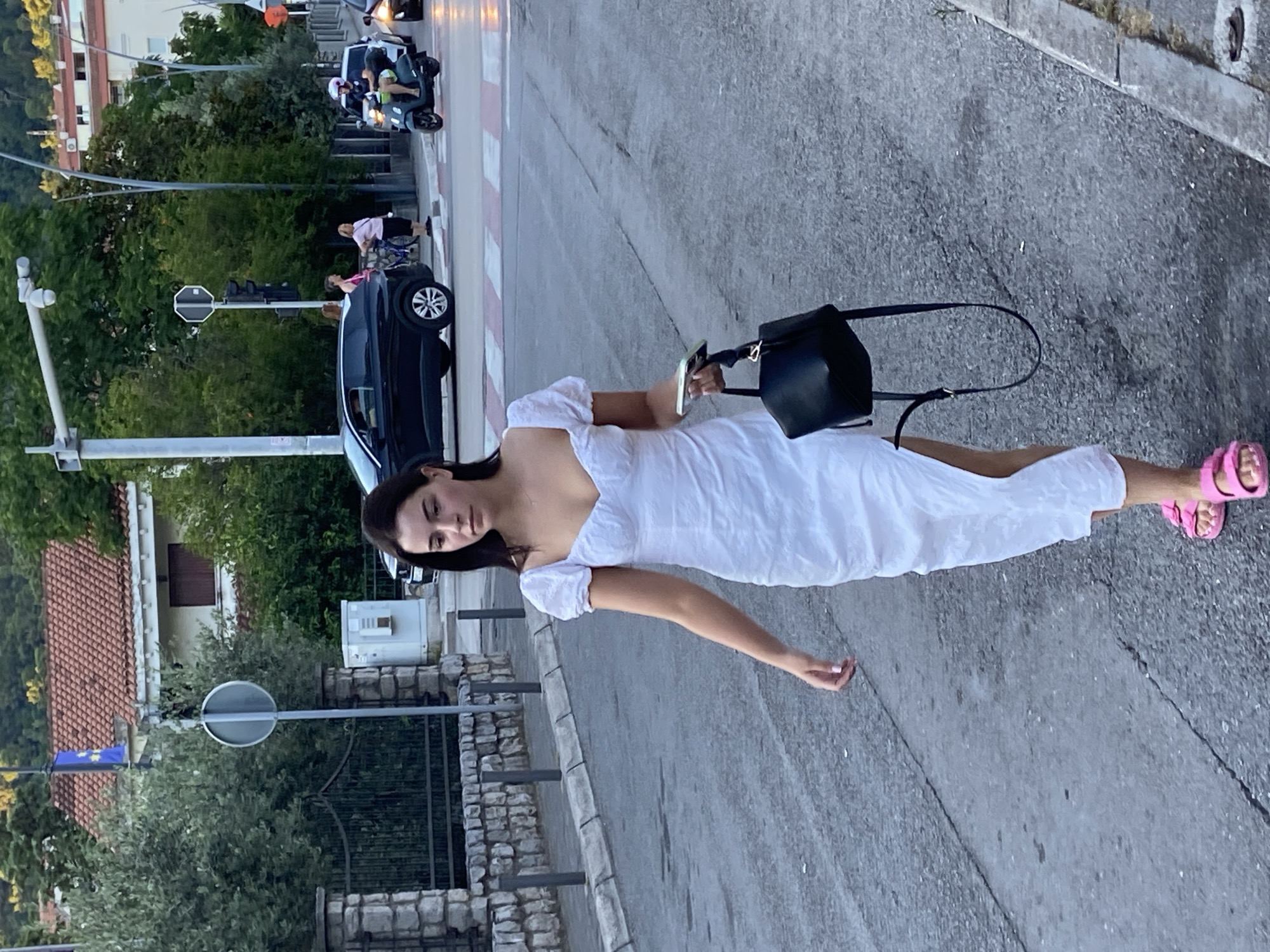 A young woman with dark hair, wearing a white linen dress and pink sandals, walks across a street in Budva, Montenegro, holding a smartphone and a black handbag. In the background, a black SUV is stopped at an intersection, and a scooter rider is passing. The street is paved with asphalt, and there are buildings with red-tiled roofs and lush greenery visible. A stone wall and fence line the left side of the street. Traffic lights and street lamps are also present.