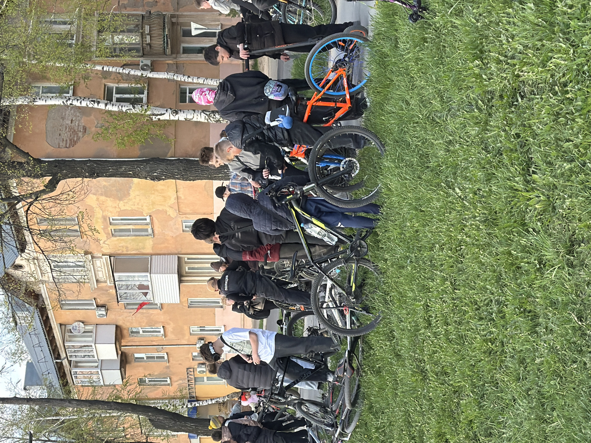 A group of people stands with their bicycles on a grassy area in Votkinsk, Russia, near a peach-colored building with white-framed windows and birch trees. The people are a mix of young adults and teens, mostly dressed in dark clothing, some with backpacks. The bicycles are various types, including mountain bikes and scooters. The building shows signs of wear and tear, with patches of exposed brick. The grassy area is lush and green, suggesting a park or public space. The setting appears to be an urban environment. Some of the people are looking at their phones. The scene suggests a gathering or meeting. A birch tree is on the left side of the building.