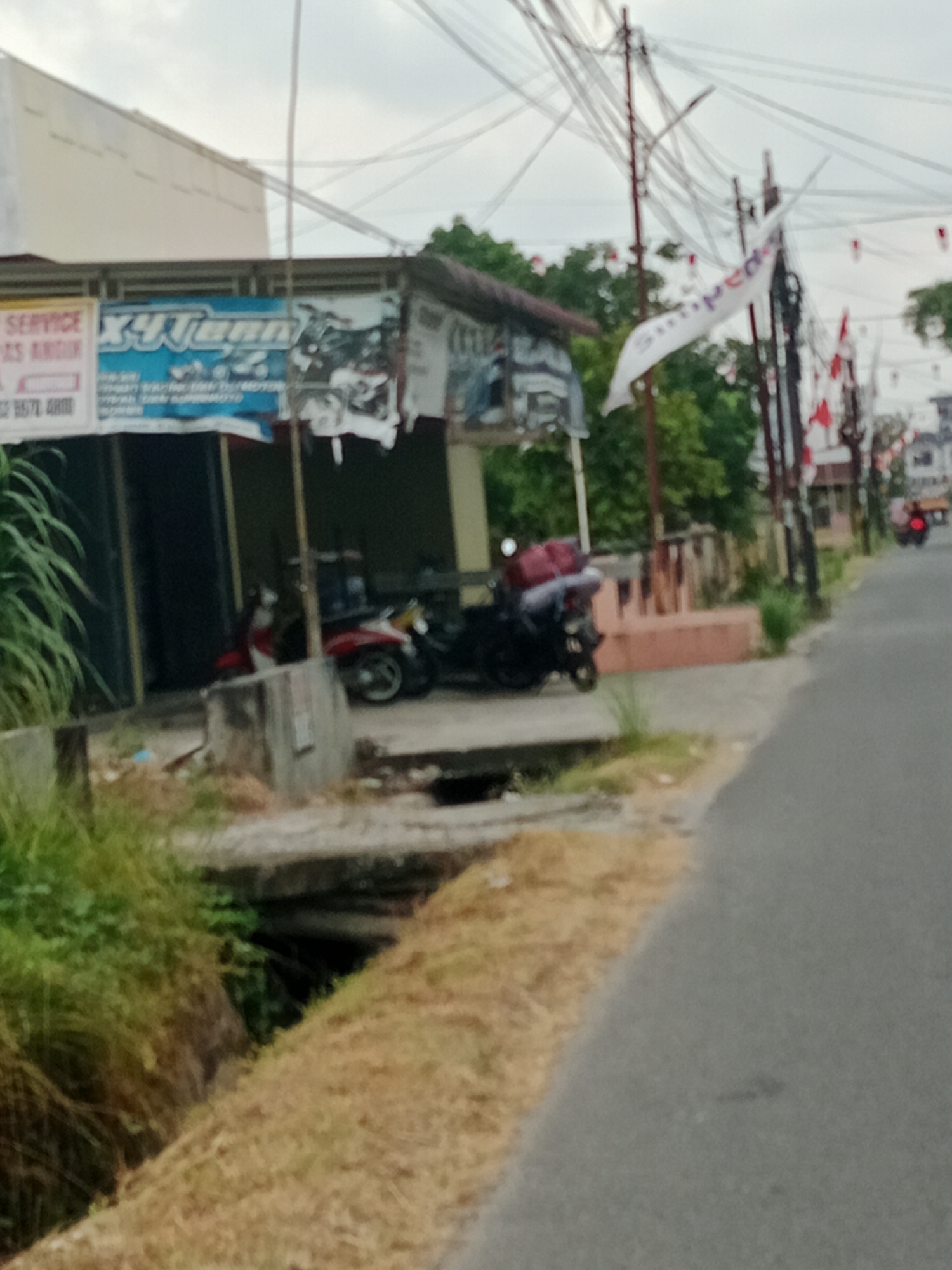 The image shows a street scene in the City of Binjai, Indonesia, with a focus on a small building that appears to be a motorcycle repair shop or a similar business. The building has a sign that reads "X4 Tenn" in blue and white lettering, with an image of a motorcycle on it. Below this sign, there are other signs, one of which is partially visible and reads "SERVICE" and "AS ARDI." There are several motorcycles parked in front of the building, some of which are covered with red tarps. A ditch filled with water and overgrown with vegetation runs alongside the road in front of the building. A paved road is visible on the right side of the image, with a few utility poles and wires overhead. Red and white flags are hung on some of the utility poles, suggesting a celebration or event. In the background, there are more buildings and trees. The sky is overcast.