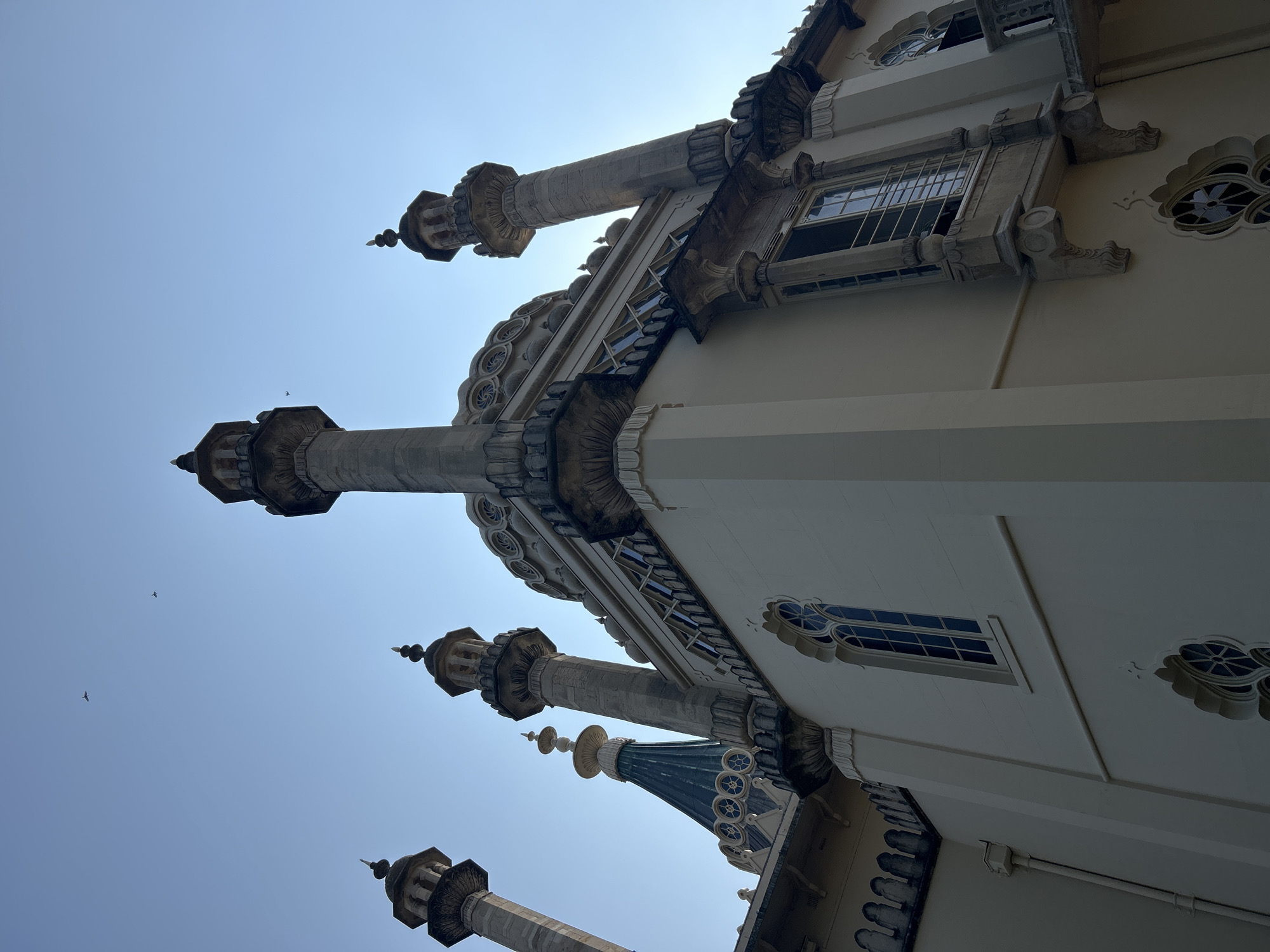 The image captures the exterior of the Royal Pavilion in Brighton, United Kingdom, viewed from a low angle against a clear, bright blue sky. The foreground features the ornate, pale cream-colored walls of the pavilion, characterized by intricate architectural details. Several minarets, with their distinctive conical tops and decorative elements, rise prominently from the structure, silhouetted against the sky. One particularly striking element is a green, conical roof with decorative circular patterns, likely a dome or spire, visible behind the minarets. Tall, arched windows with dark frames and mullions punctuate the walls, some featuring barred coverings. The building exhibits a blend of architectural styles, with Islamic and Indian influences evident in its design. In the upper portion of the sky, two small, dark silhouettes of birds are visible, adding a sense of scale and life to the serene atmosphere. The lighting suggests it is daytime, with the sun casting some shadows and highlighting the building's textures.