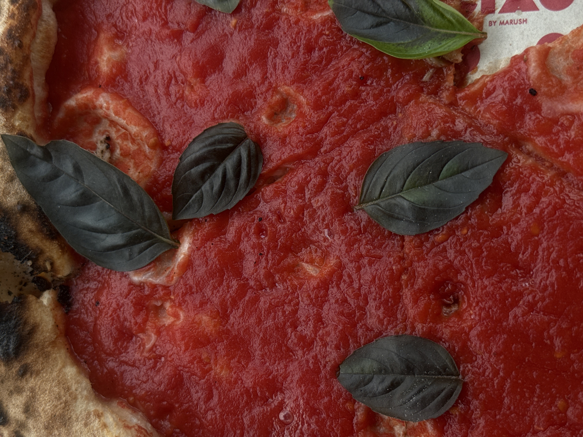 Close-up shot of a pizza with a tomato sauce base and basil leaves on top. The pizza crust is visible on the left and upper right edges, showing some charring. The pizza appears to be served on a cardboard box with the inscription "BY MARUSH" visible in the upper right corner. There is no indication of a specific location in Yerevan, Armenia. There are no people or actions visible.