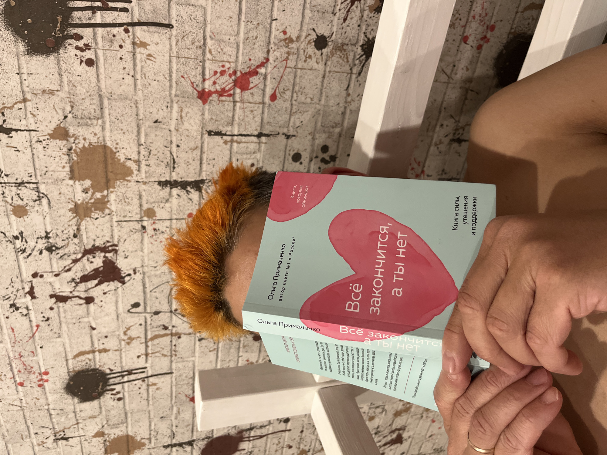 A woman with orange hair is lying on a white wooden bench indoors. She is holding a book titled "Всё закончится, а ты нет" by Olga Primachenko in front of her face. The book's cover has a pink heart on it. The wall behind the woman is painted with white brick pattern and has splatters of brown, black, and red paint. The book is described as a "Book of Power, comfort, and support". There is a gold wedding band on the woman's left hand. The author of the book is described as an author of book #1 in Russia. The author herself is described as a creator of Super bestseller book "To myself."